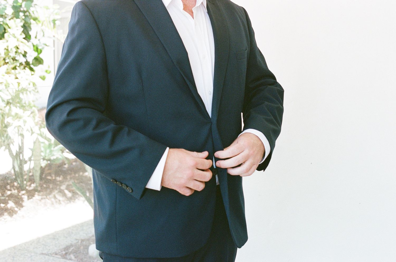 Groom in navy suit buttoning jacket before tropical garden wedding ceremony.