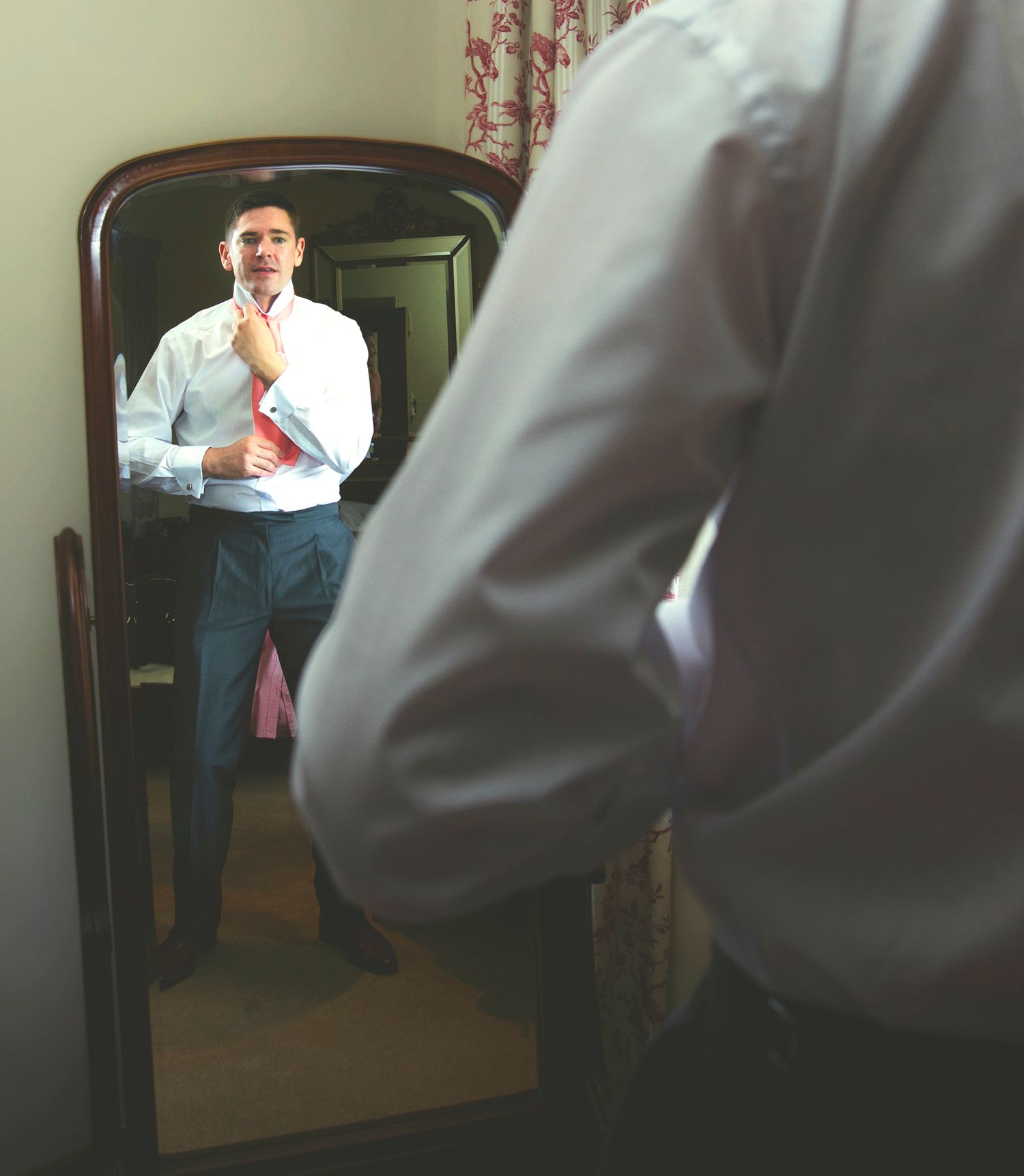 Groom adjusting his coral pink tie in a full-length wooden mirror during morning preparations.