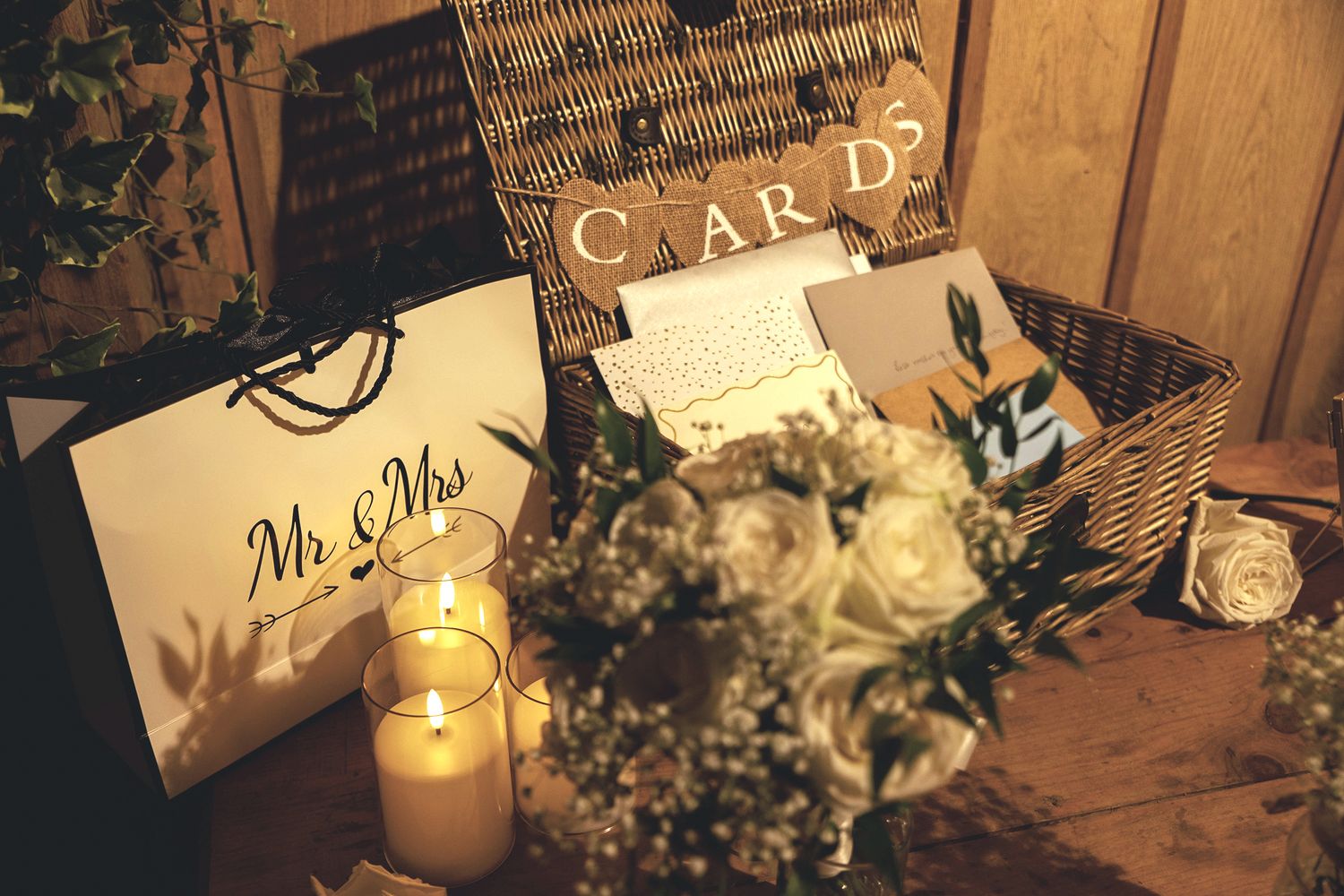 Wedding reception display table featuring a wicker card basket, burning candles, and a white Mr and Mrs gift bag.