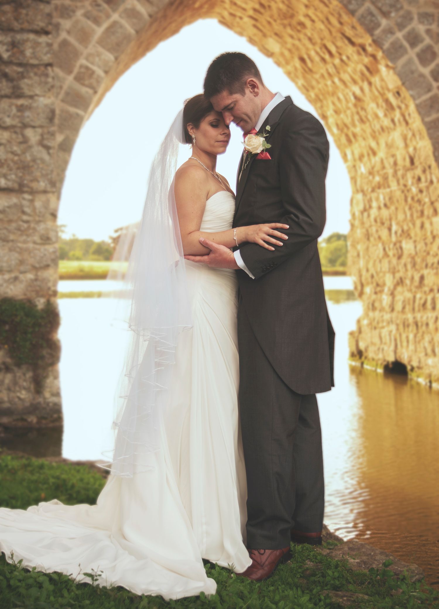 Bride in a strapless wedding dress and groom in a morning coat embracing under a stone archway by a river.