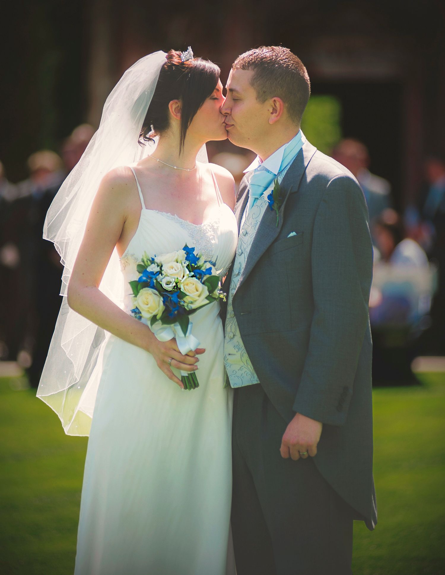 Bride and groom kissing outdoors in sunlight, featuring a blue and white rose bouquet and grey morning suit.