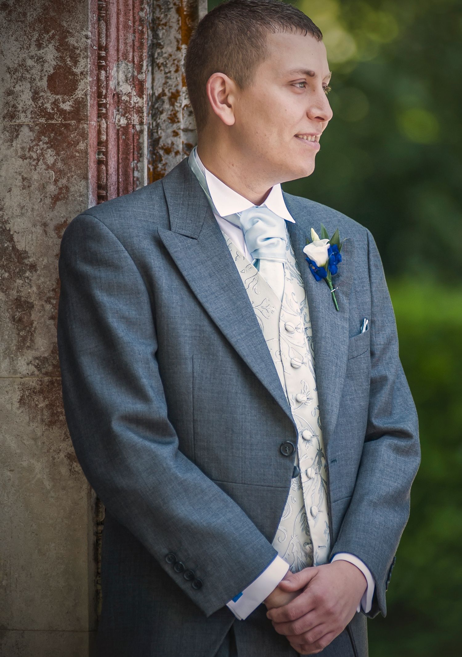 Groom profile portrait in a grey morning suit, patterned waistcoat, and light blue cravat, leaning against a stone wall.