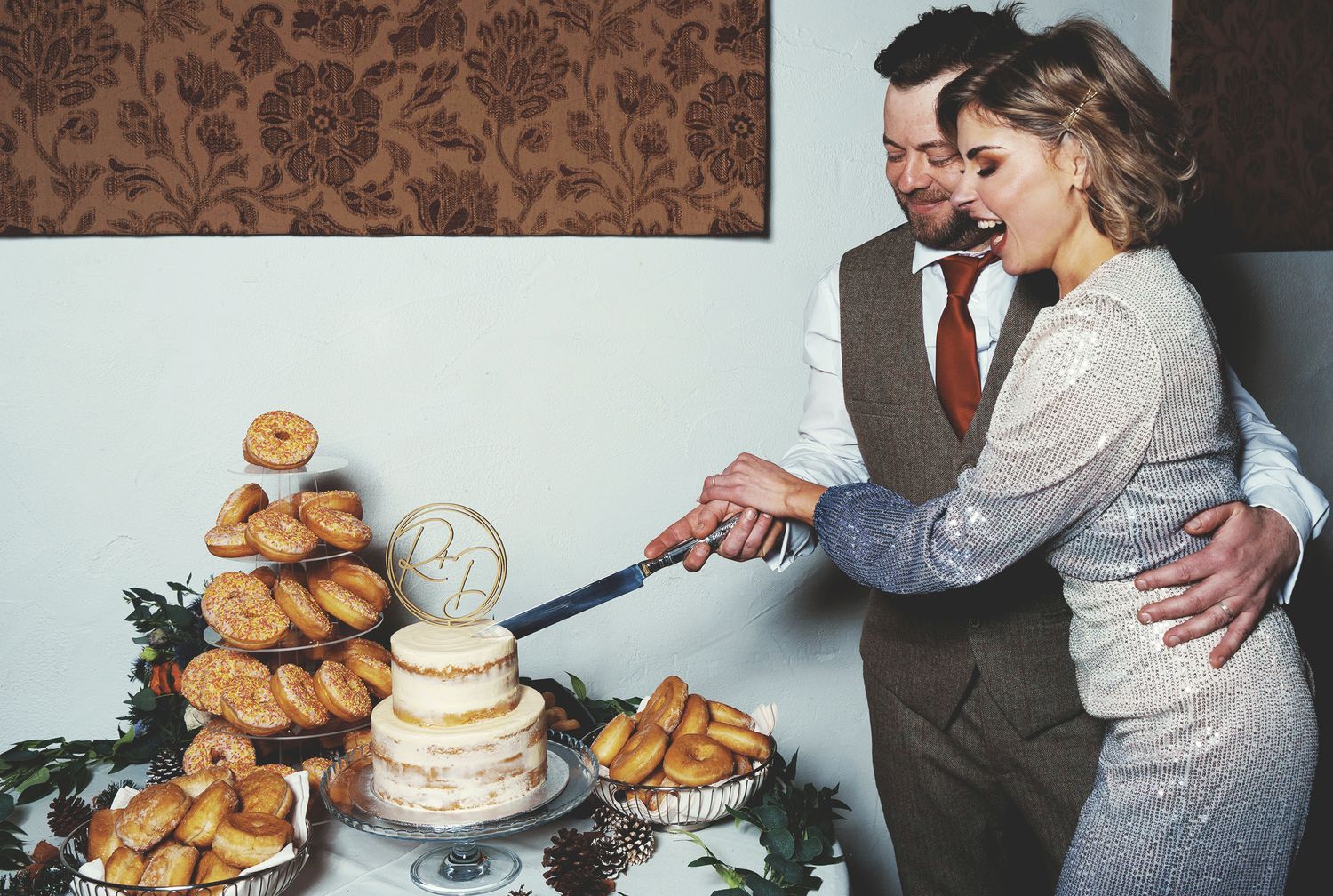 Bride in a sequin jump suit and groom in a tweed waistcoat cutting a small naked cake surrounded by donuts.