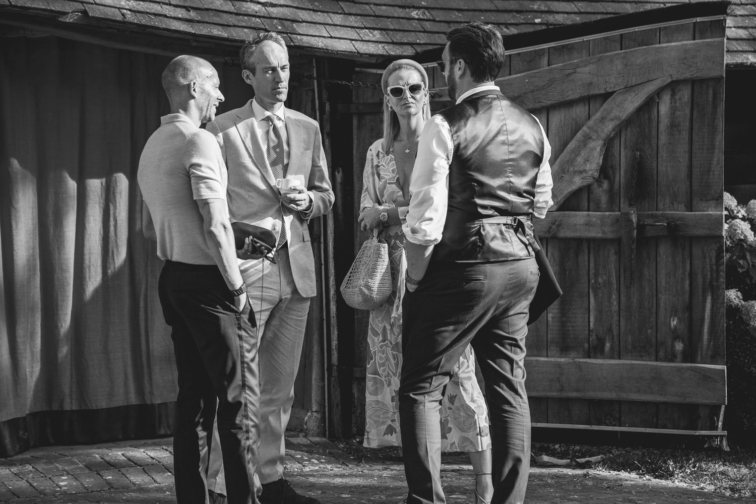 Black and white candid photograph of wedding guests chatting outside a rustic barn venue in afternoon sunlight.