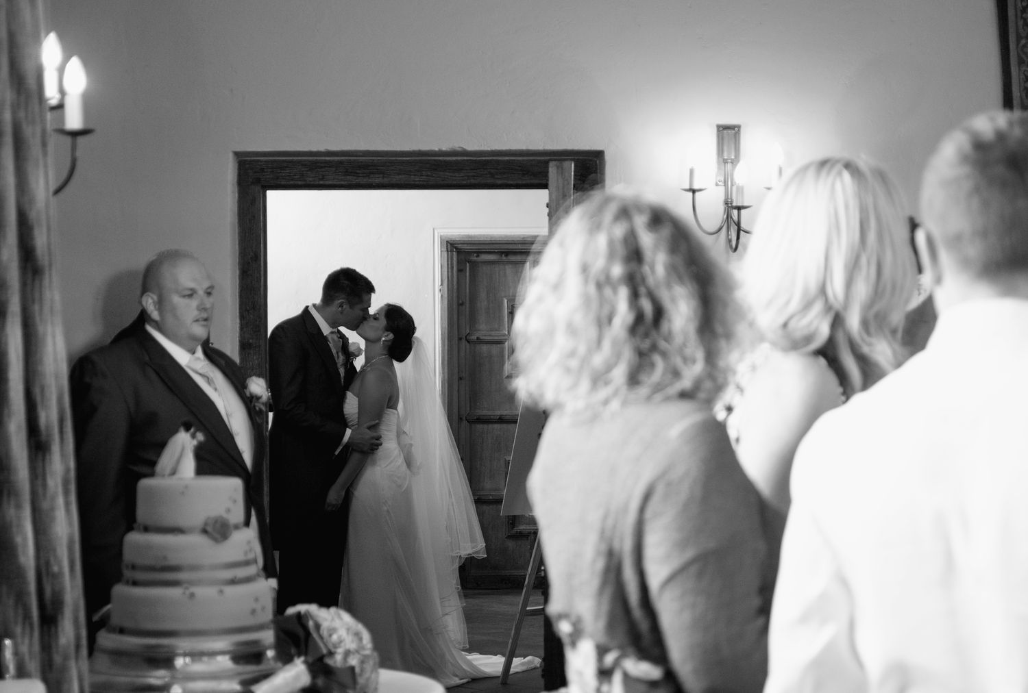 Black and white candid shot of a bride and groom kissing in a doorway during their reception, framed by guests.