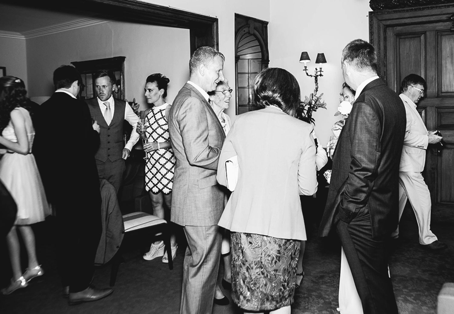 Black and white candid photograph of wedding guests socialising indoors during a drinks reception.