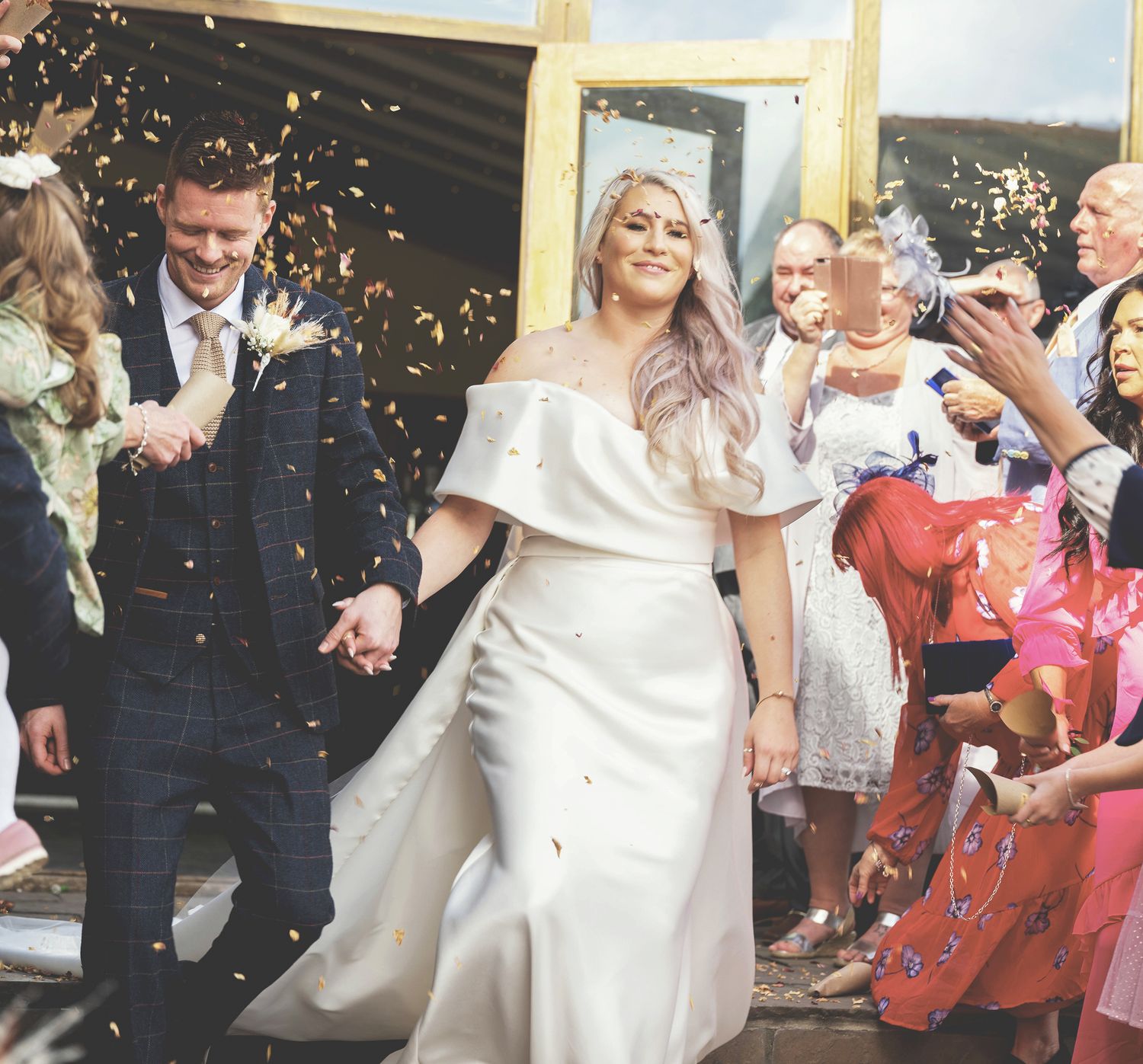 Bride and groom smiling during their confetti exit as wedding guests throw gold confetti outside a venue.