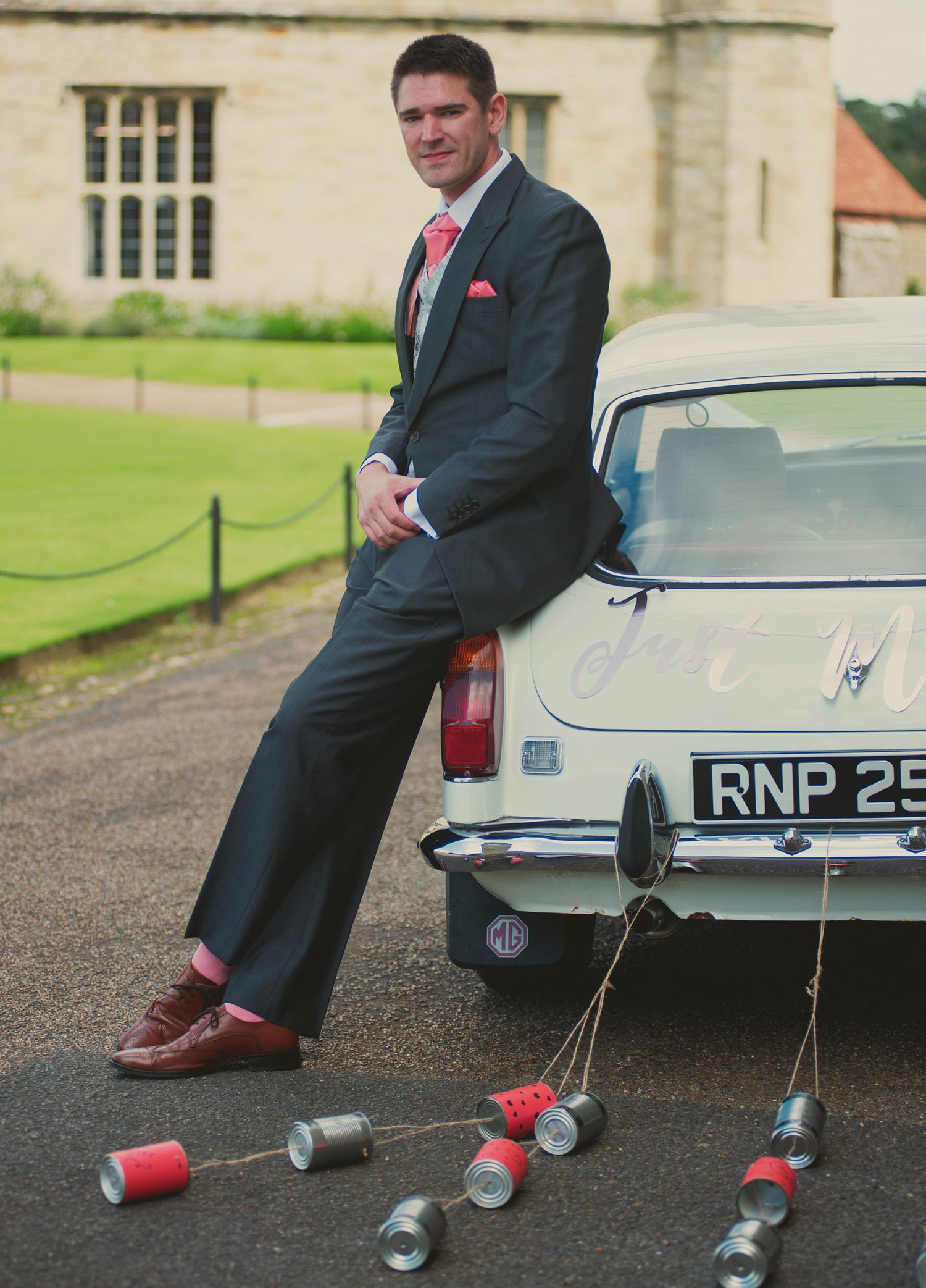 Groom leaning against a white vintage wedding car trailing tin cans, with a country house venue in the background.