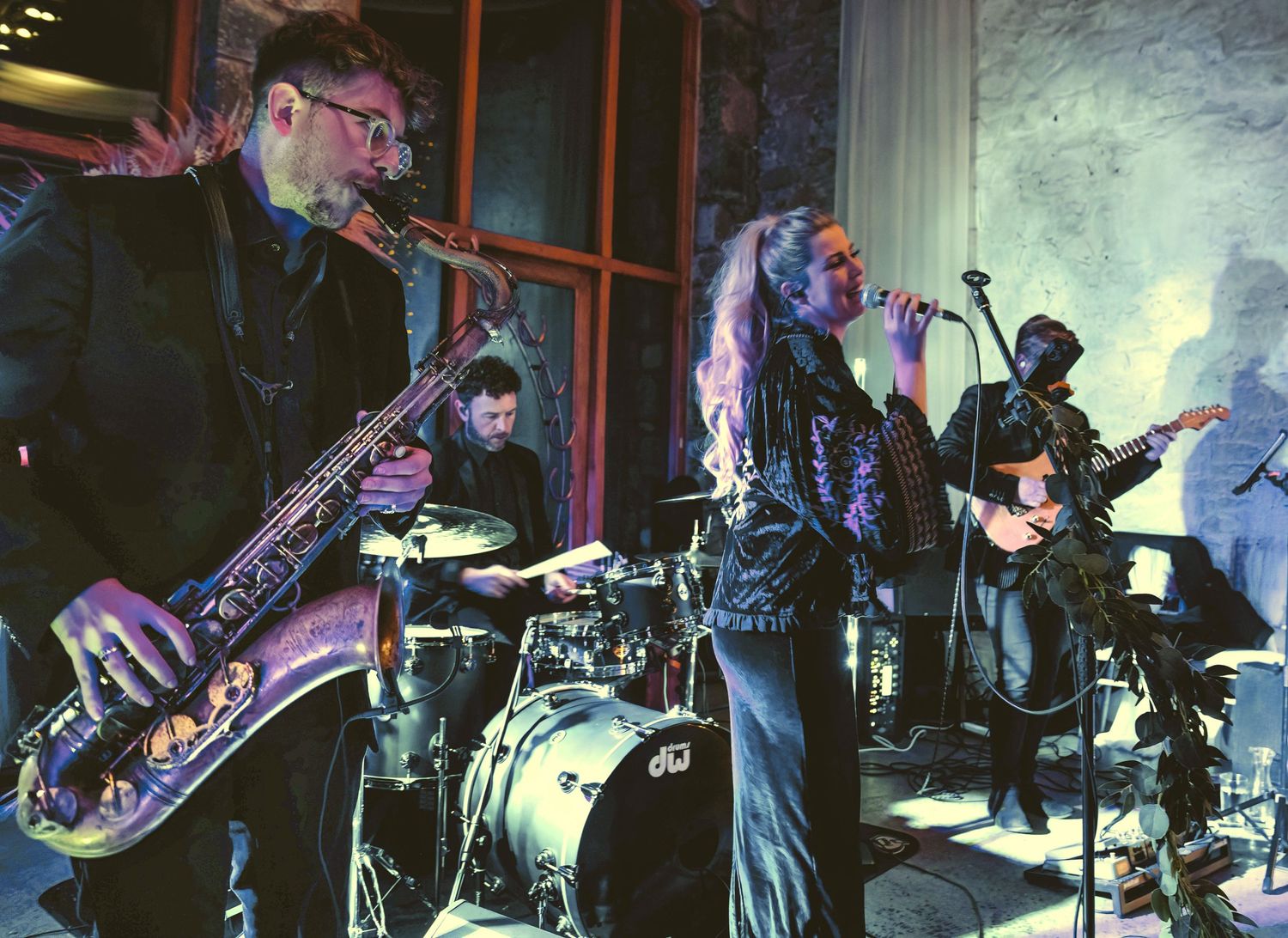 Live wedding band performing at an evening reception with a female singer, a saxophonist, and a drummer under stage lights.