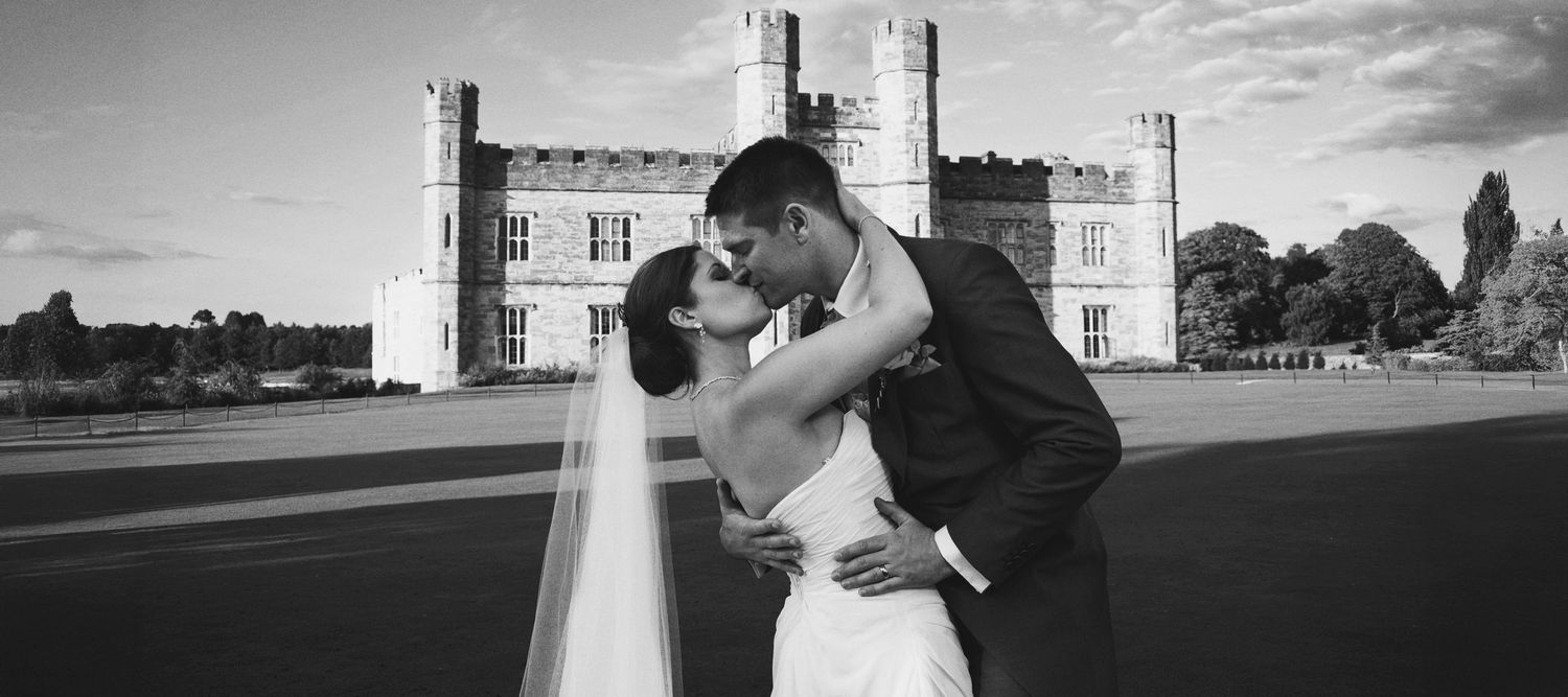 Black and white photograph of a bride and groom kissing, with Leeds Castle in the background.