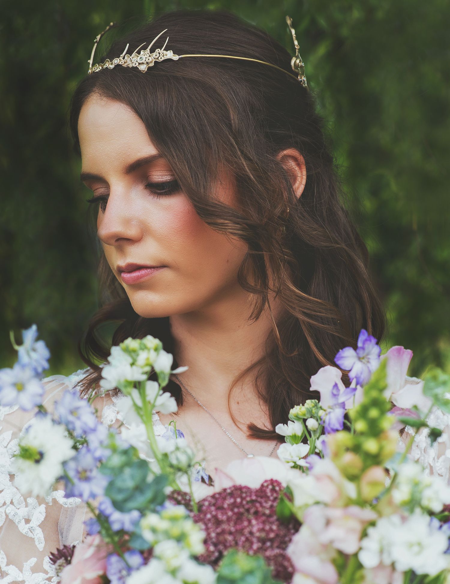 Close-up portrait of a bride looking down, wearing a gold tiara crown with purple and white flowers in the foreground.