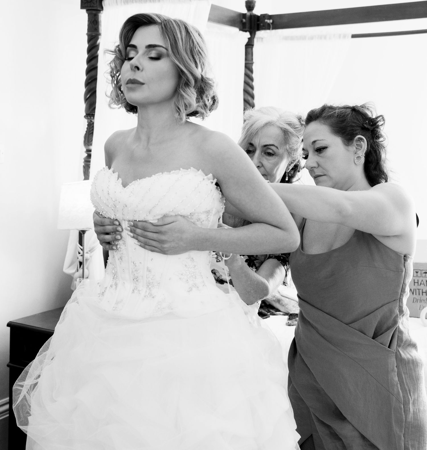 Black and white candid photograph of a bride having her strapless wedding dress adjusted during morning preparations.