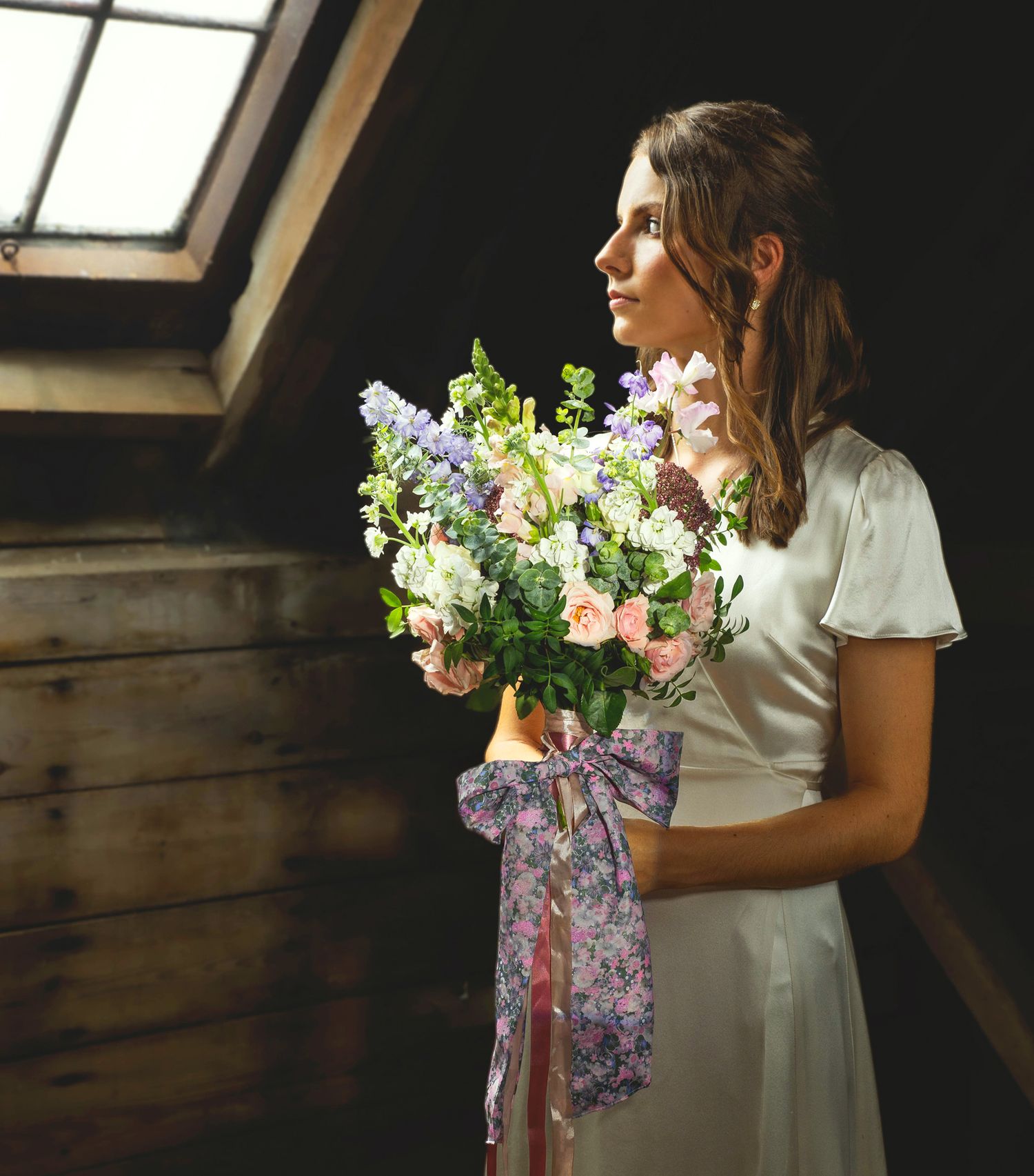 Bride in a satin dress holding a large bouquet with a floral ribbon, standing by a wooden window in soft directional light.