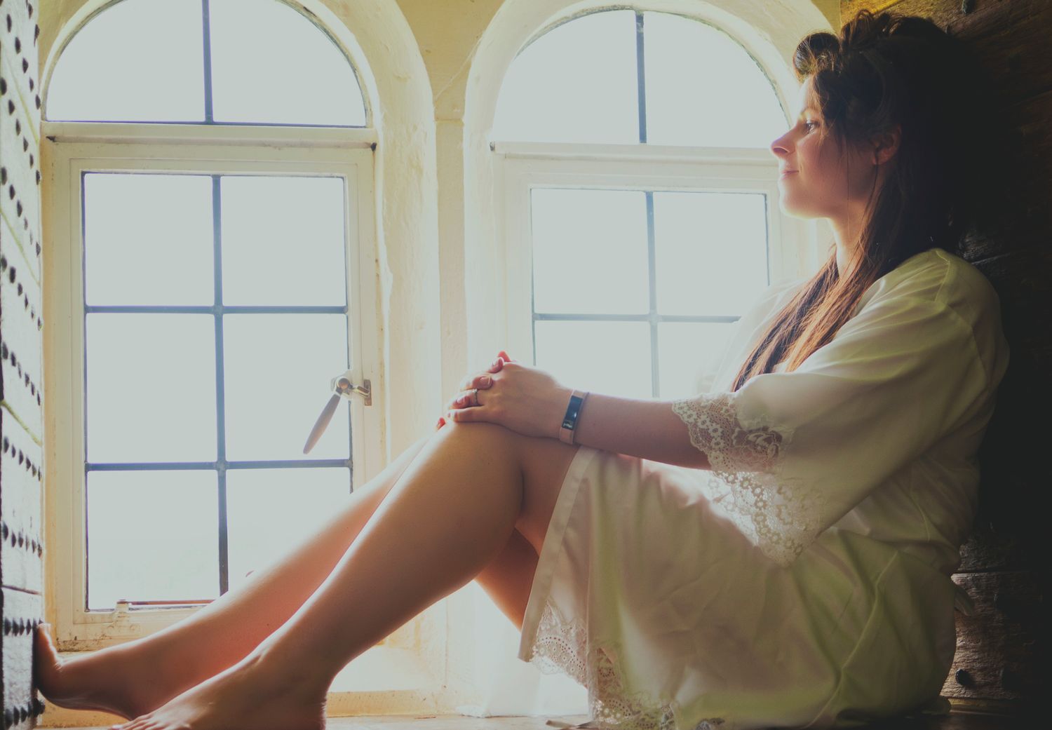 Bride wearing a lace-trimmed bridal robe sitting on a window sill during morning wedding preparations.