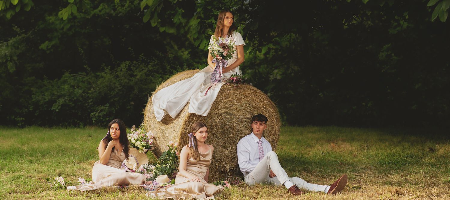 Kincora homepage banner of an editorial wedding photo of a bride sitting on a round hay bale with bridesmaids and groomsman relaxed below.