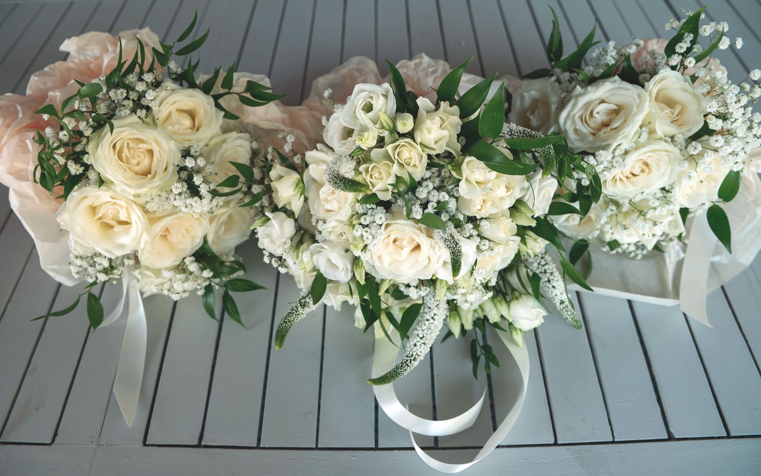 Three matching bridal bouquets with white roses, gypsophila, and green foliage resting on a grey wooden surface.