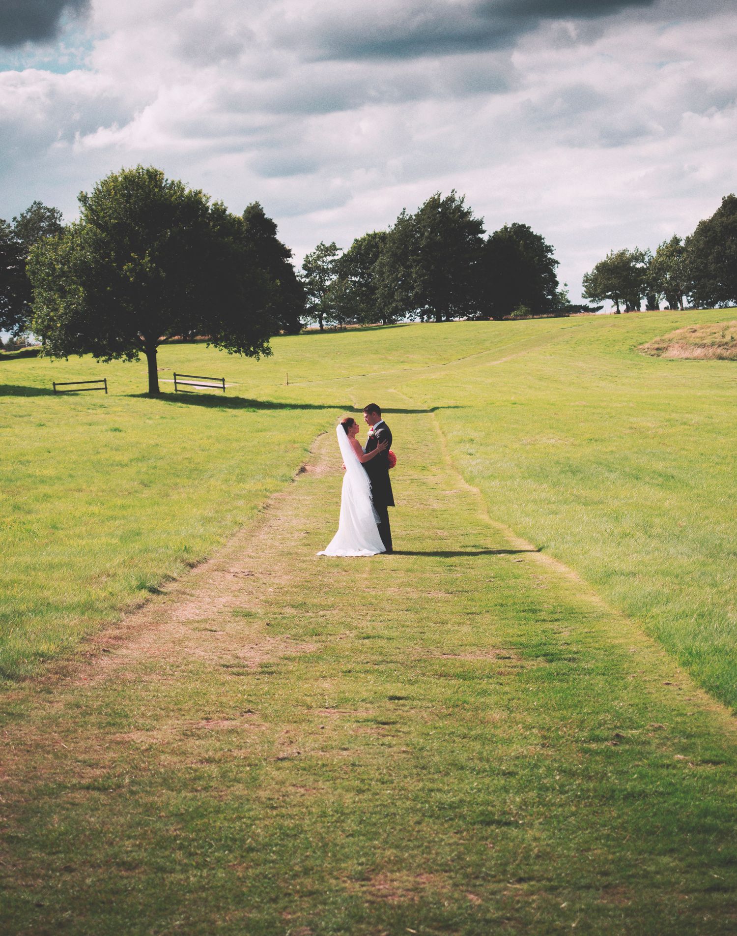 Wide landscape portrait of a bride and groom standing together on a grass path in an open countryside estate field.