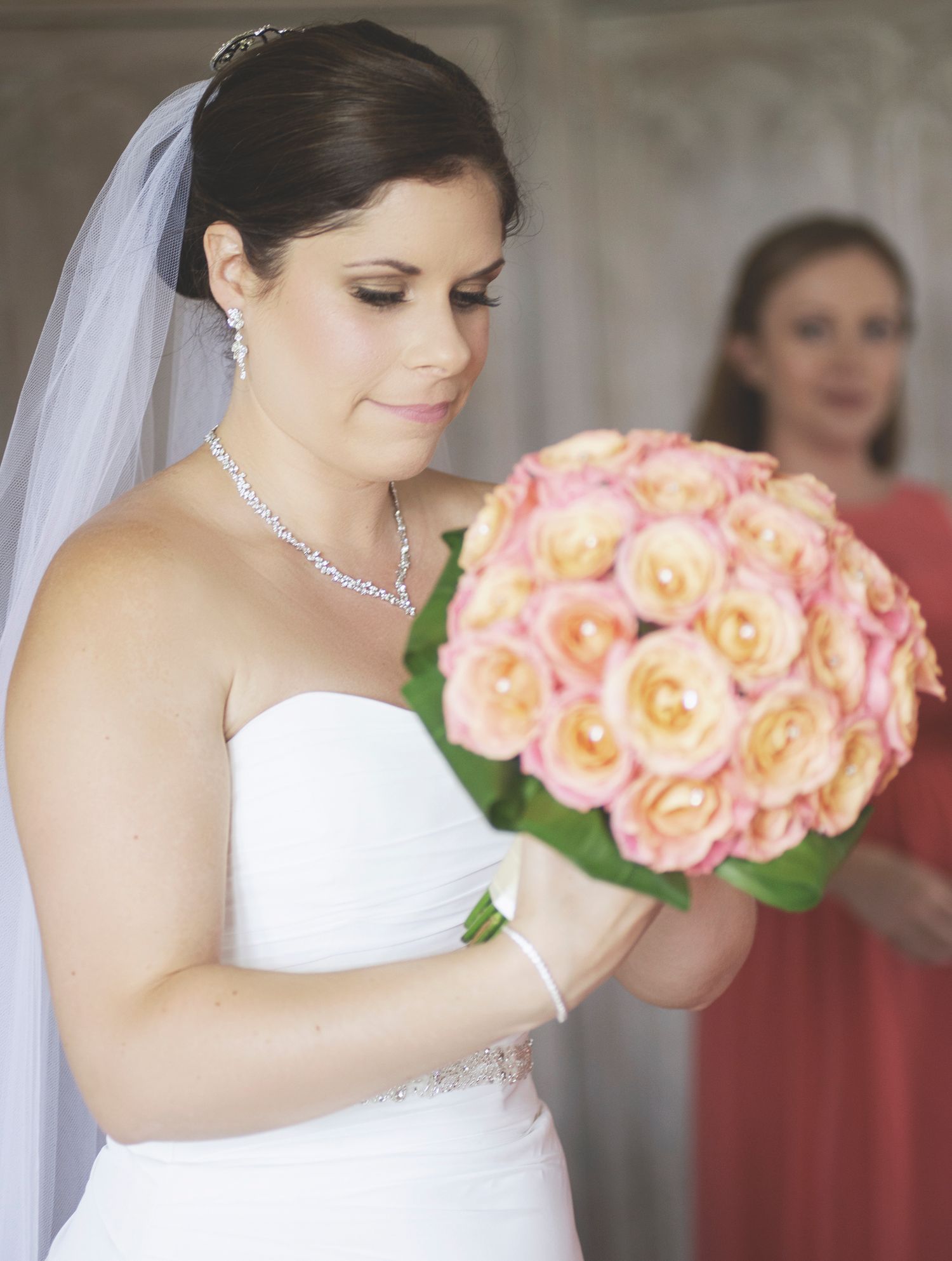 Bride looking down at a peach rose bridal bouquet during morning preparations, with a bridesmaid in the background.