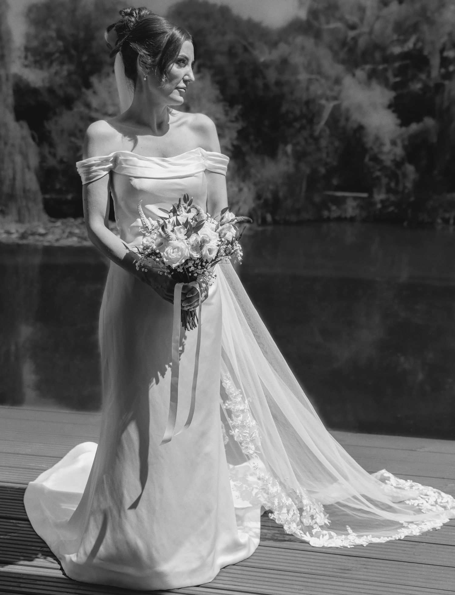 Black and white bridal portrait in an off-the-shoulder satin gown with a long lace-trimmed veil by a lake.