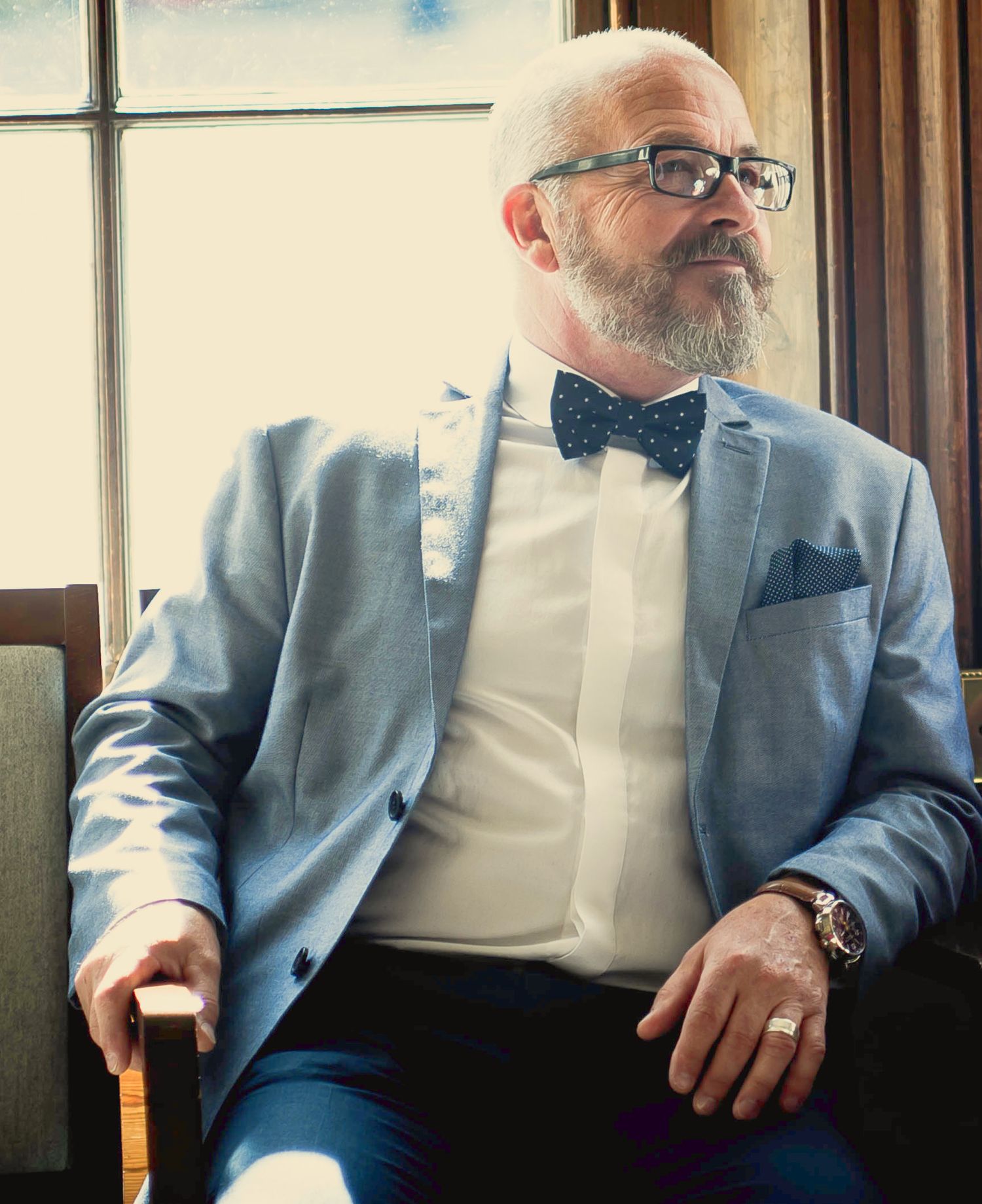 Portrait of a bearded wedding guest in a light blue blazer, patterned bow tie, and glasses sitting near a window.