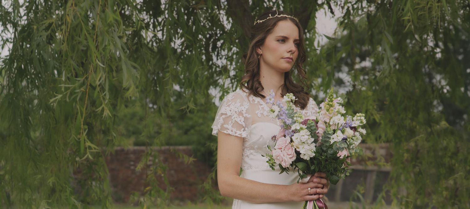 Bride in a short-sleeved lace wedding dress holding a pastel bouquet under a weeping willow tree.