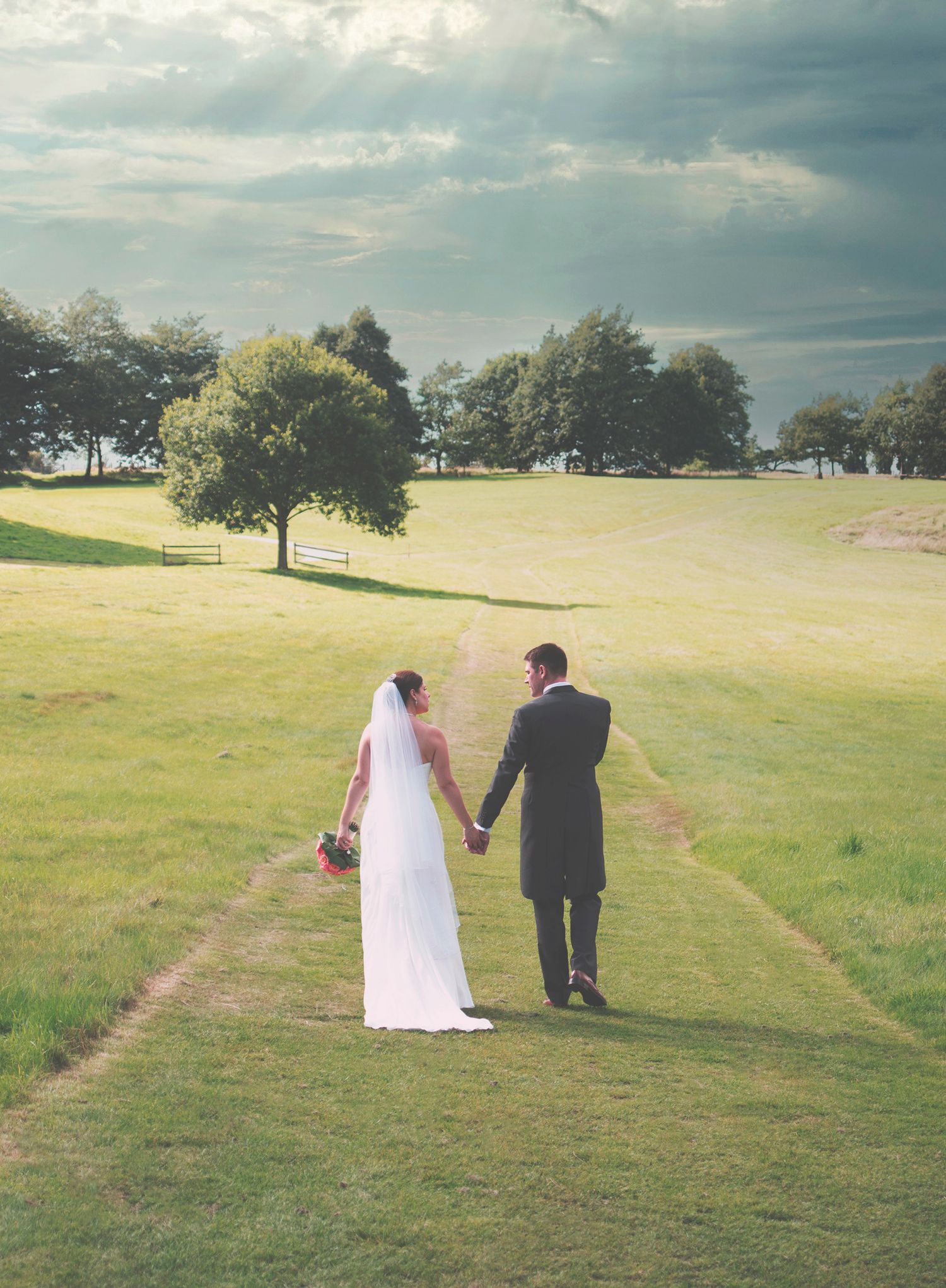 Bride and groom holding hands, seen from behind as they walk down a grass path through an open estate field.