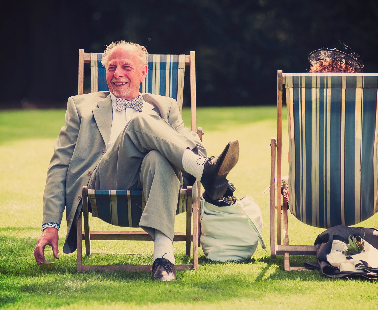 Smiling elderly wedding guest in a suit and bow tie relaxing in a striped deckchair on a garden lawn.