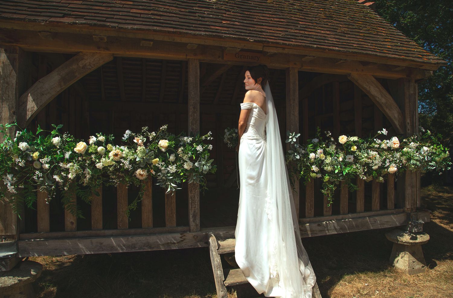 Bride in a satin gown and long veil standing inside a rustic oak gazebo decorated with white floral garlands.