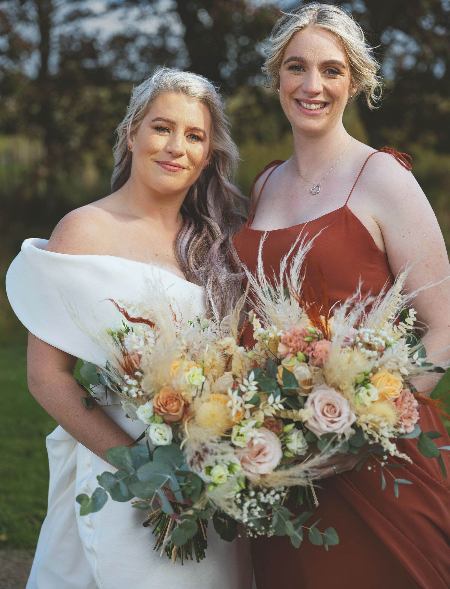 Bride in an off-the-shoulder gown standing beside her bridesmaid in a rust red dress, holding pampas grass bouquets.