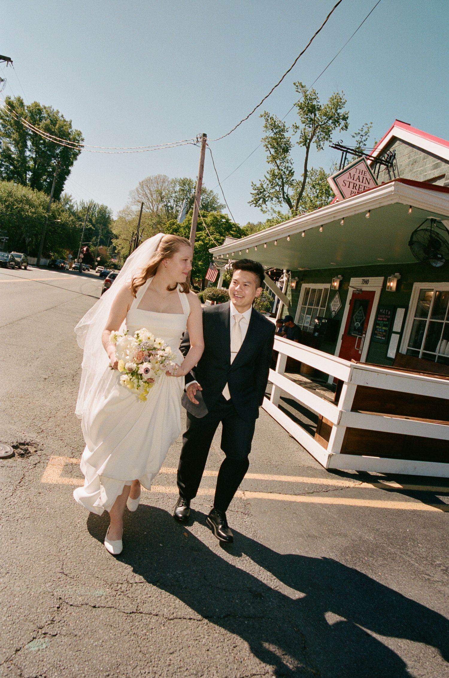 Bride and groom walking hand-in-hand on a sunny street, bride in white gown holding bouquet, groom in dark suit.
