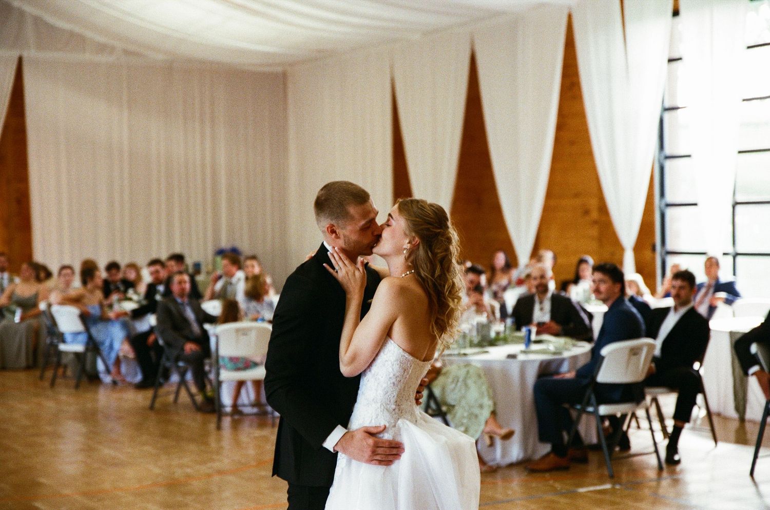 Bride and groom share first dance kiss at elegant wedding reception with guests seated at decorated tables.