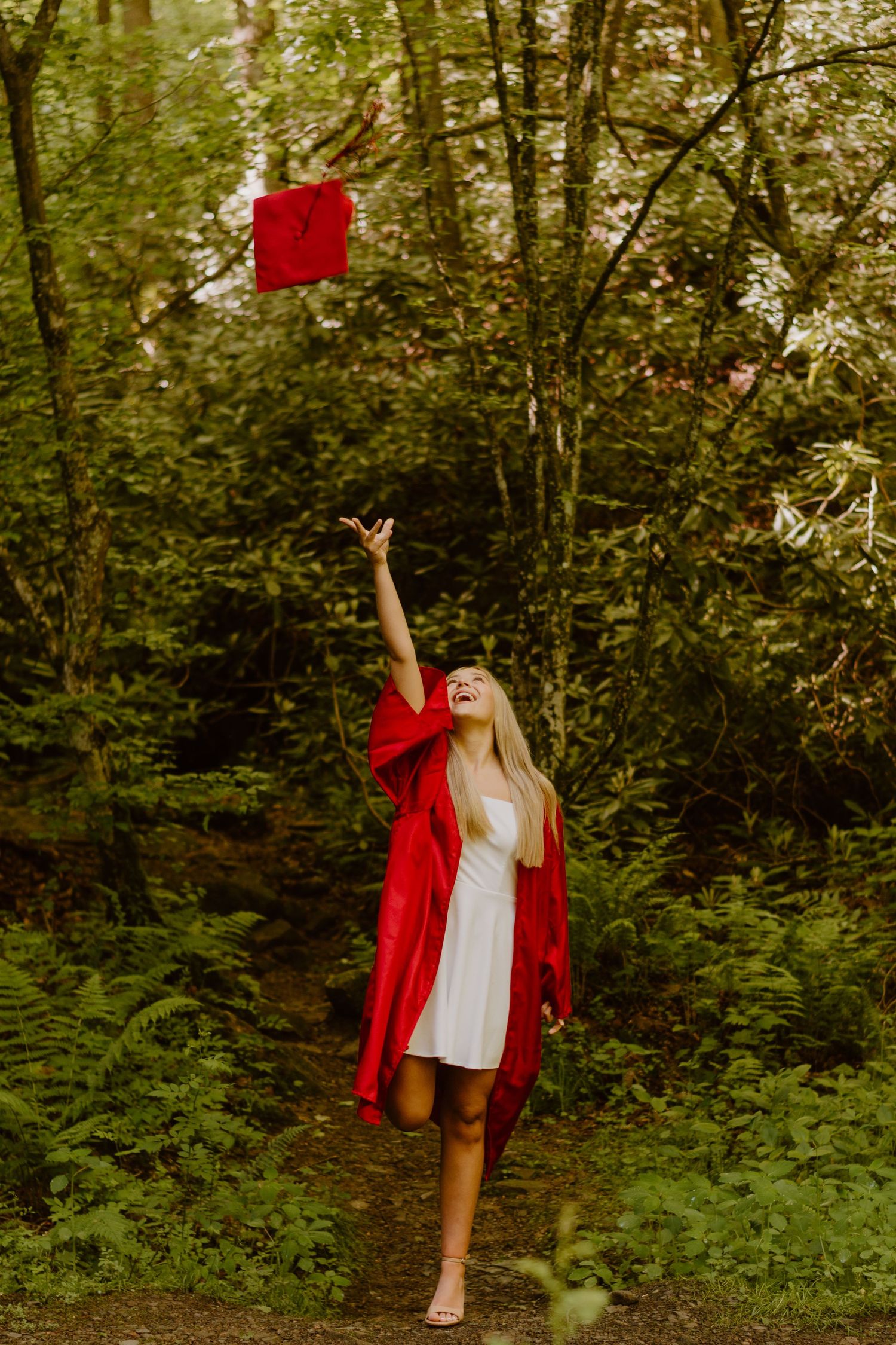 Smiling female graduate in red gown tossing cap in lush green forest setting.