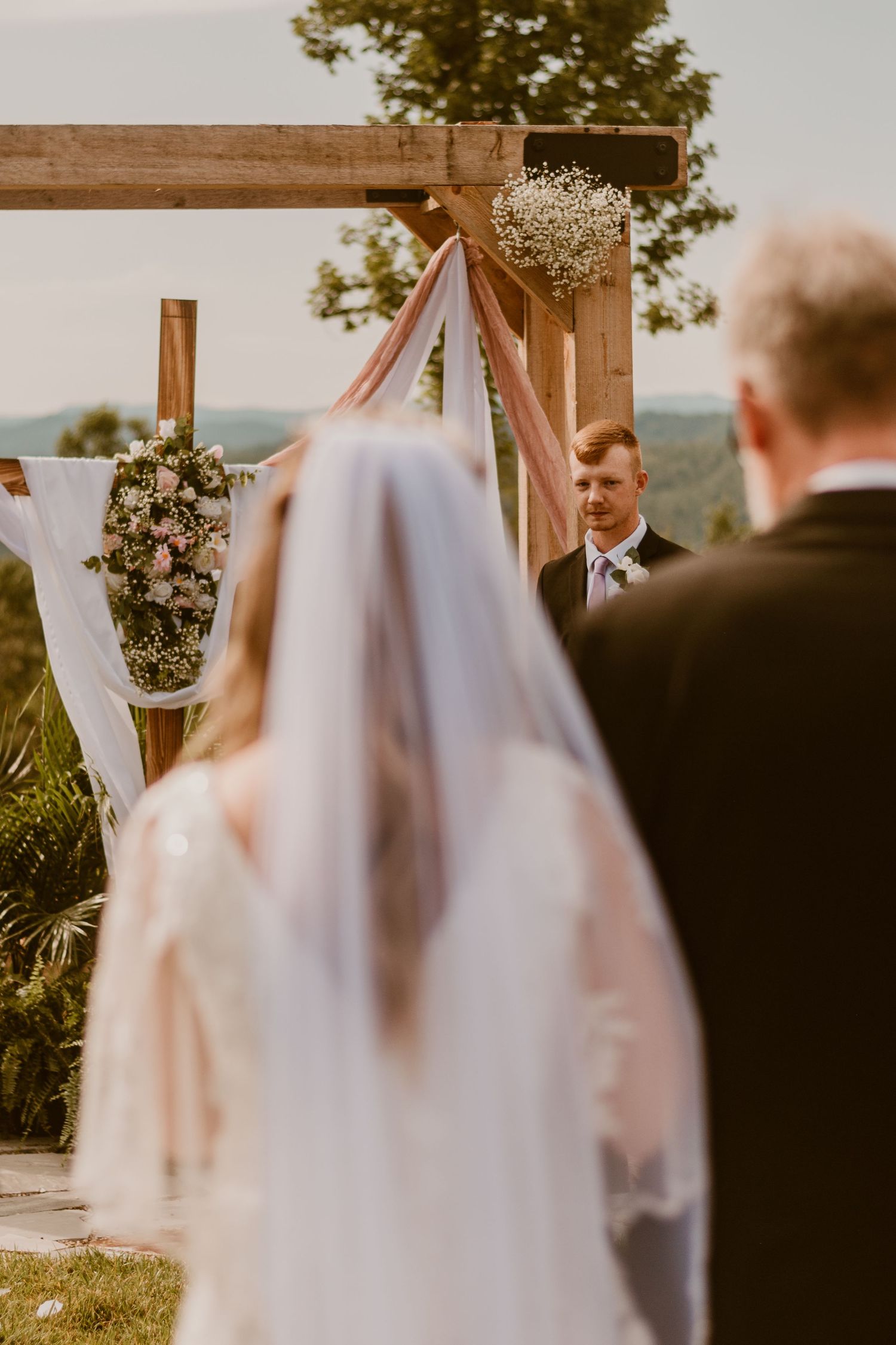 Outdoor wedding ceremony with bride in veil approaching groom at rustic wooden arch adorned with pink flowers and draping.