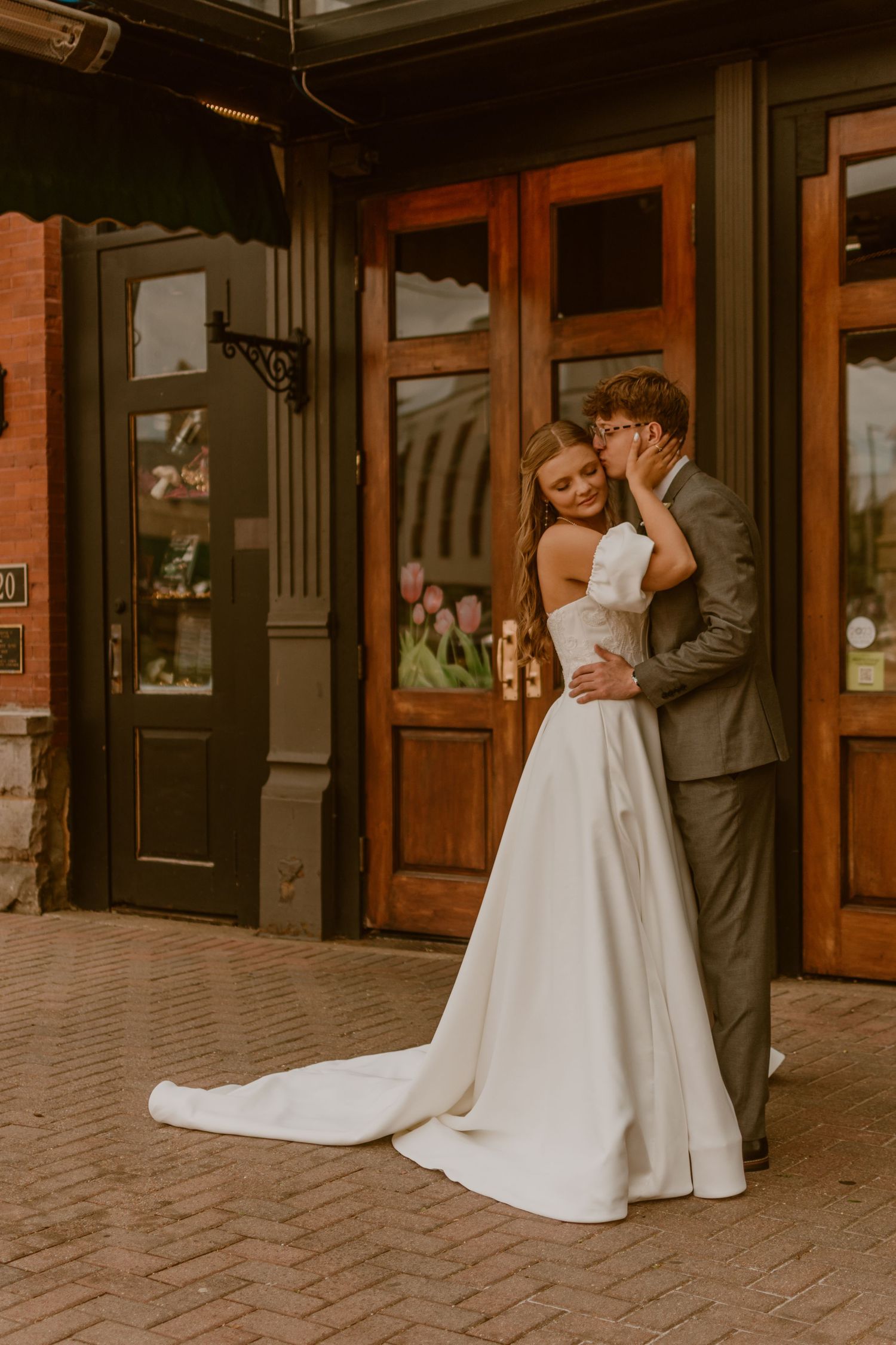 A groom in a gray suit whispers to his bride in a white gown with train, standing before wooden doors.