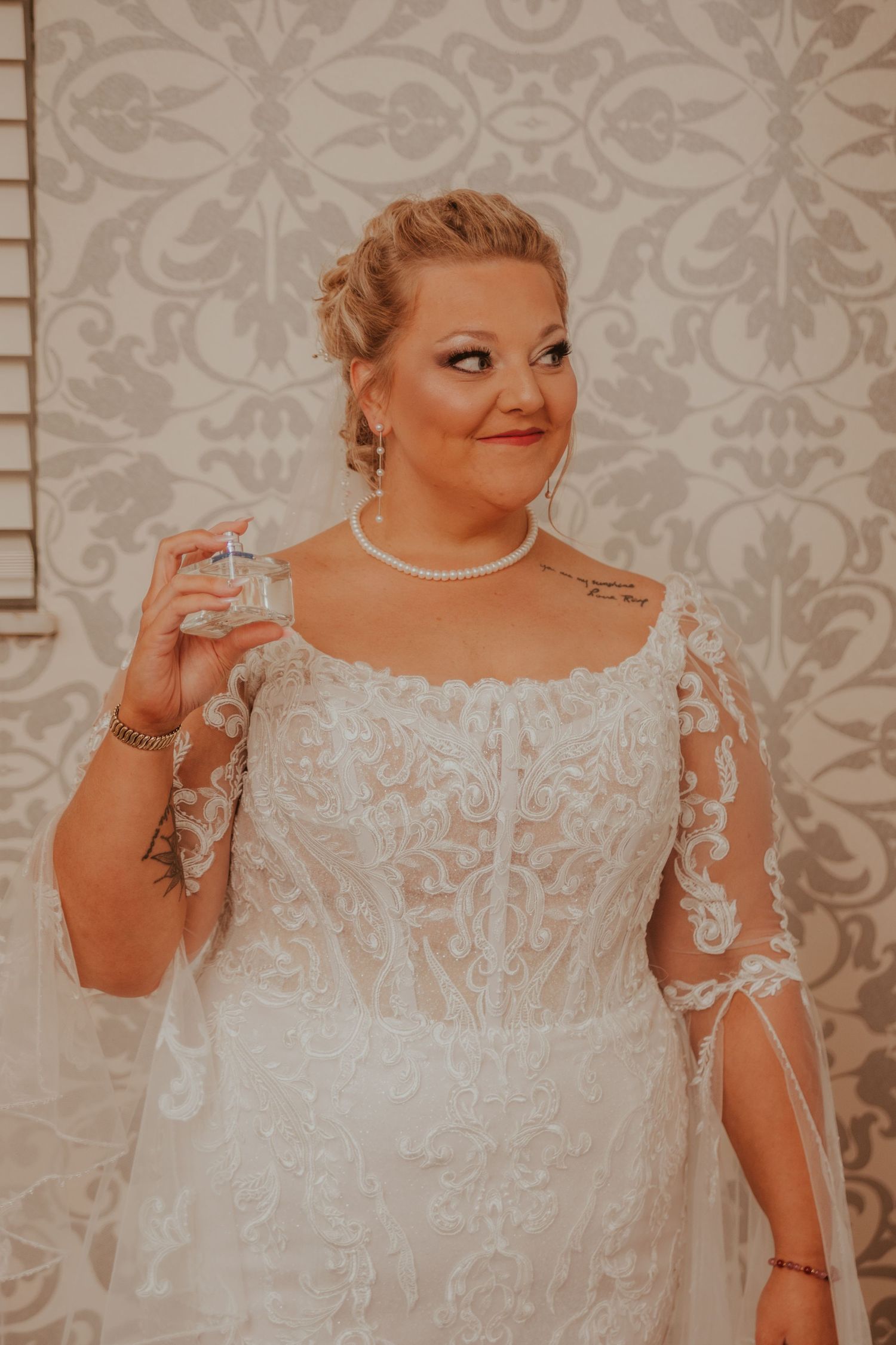 Beautiful lace wedding dress with intricate details and sheer sleeves against a damask wallpaper background.