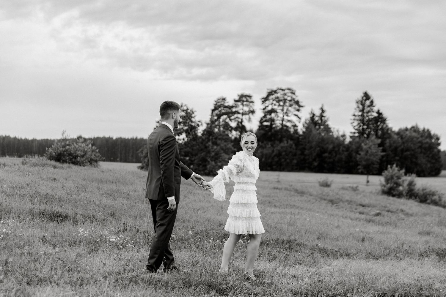 Hääpari kävelee iltapotreteissa kauniilla niityllä käsikädessä mustavalkokuvassa