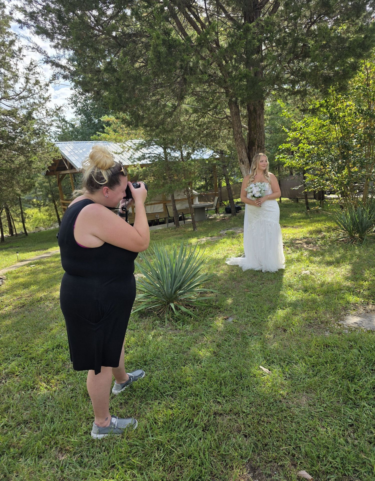 Photographer capturing a bride in a white gown holding flowers outdoors among trees and green grass.