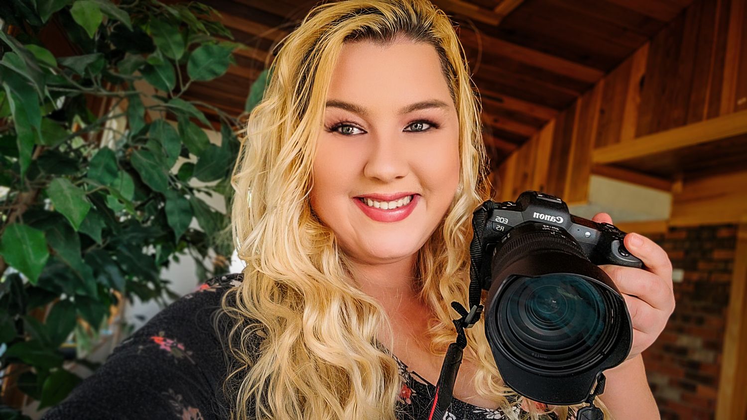 Professional female photographer with blonde hair smiles while holding a DSLR camera in a rustic wooden setting indoors with green plants.