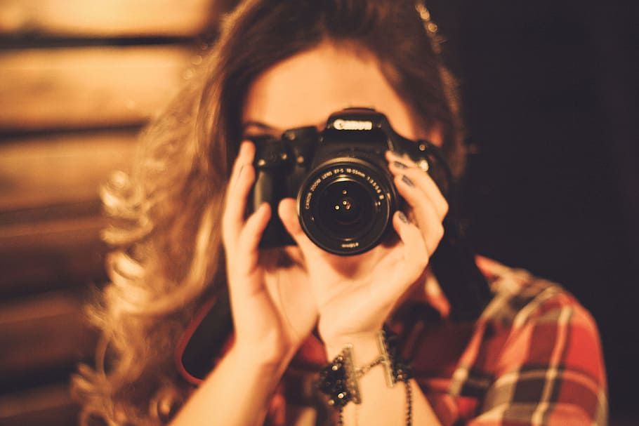 Young woman with curly hair holding a Canon DSLR camera up to her eye, wearing a plaid shirt.