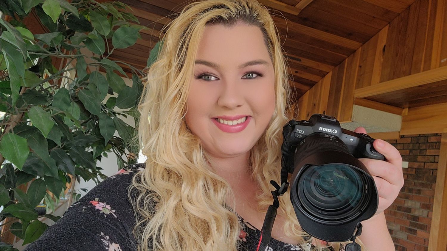 Smiling woman with long wavy blonde hair holds a black mirrorless camera on a wooden porch with green plants nearby.