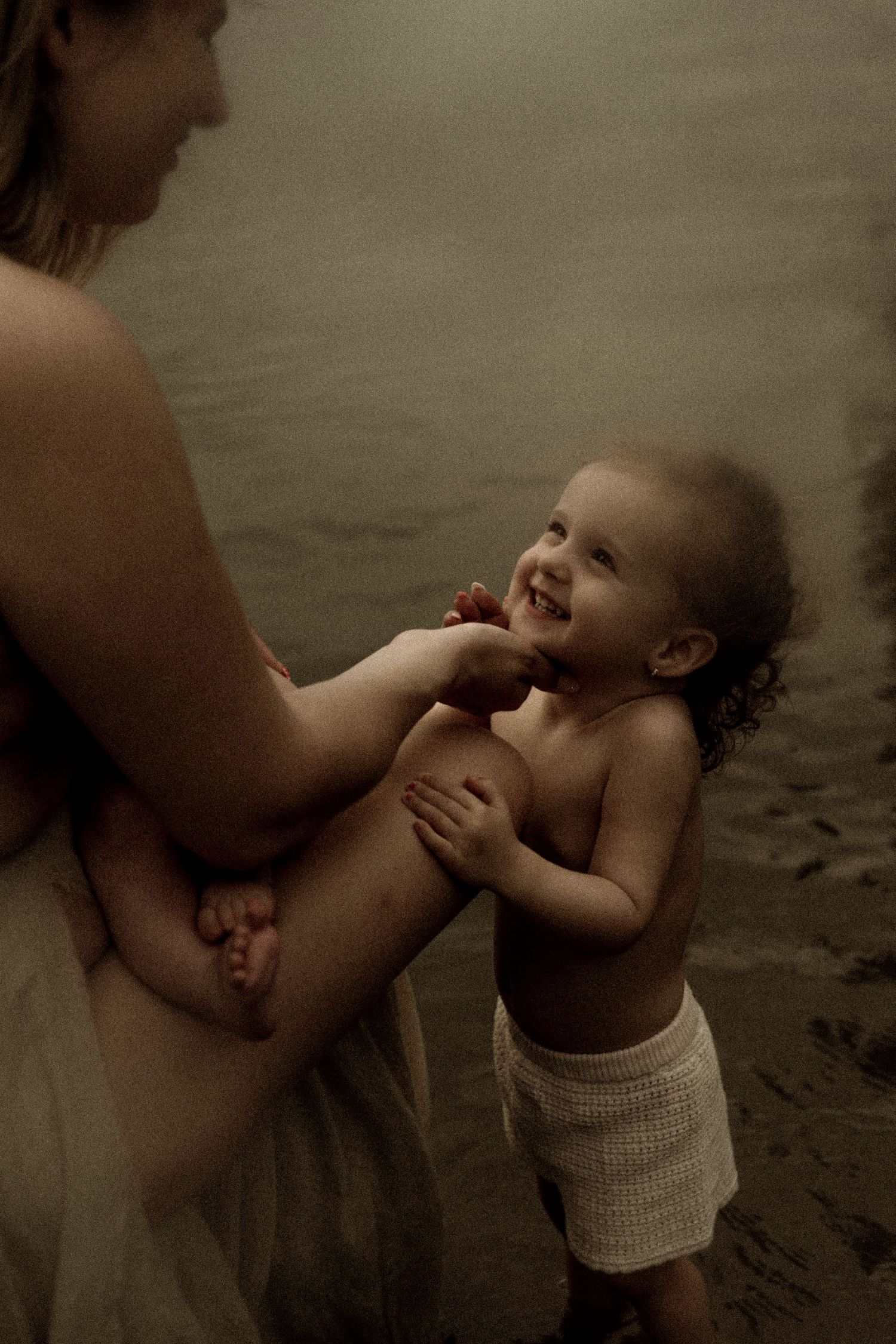 A playful moment between parent and toddler captured in sepia tones at the water's edge.