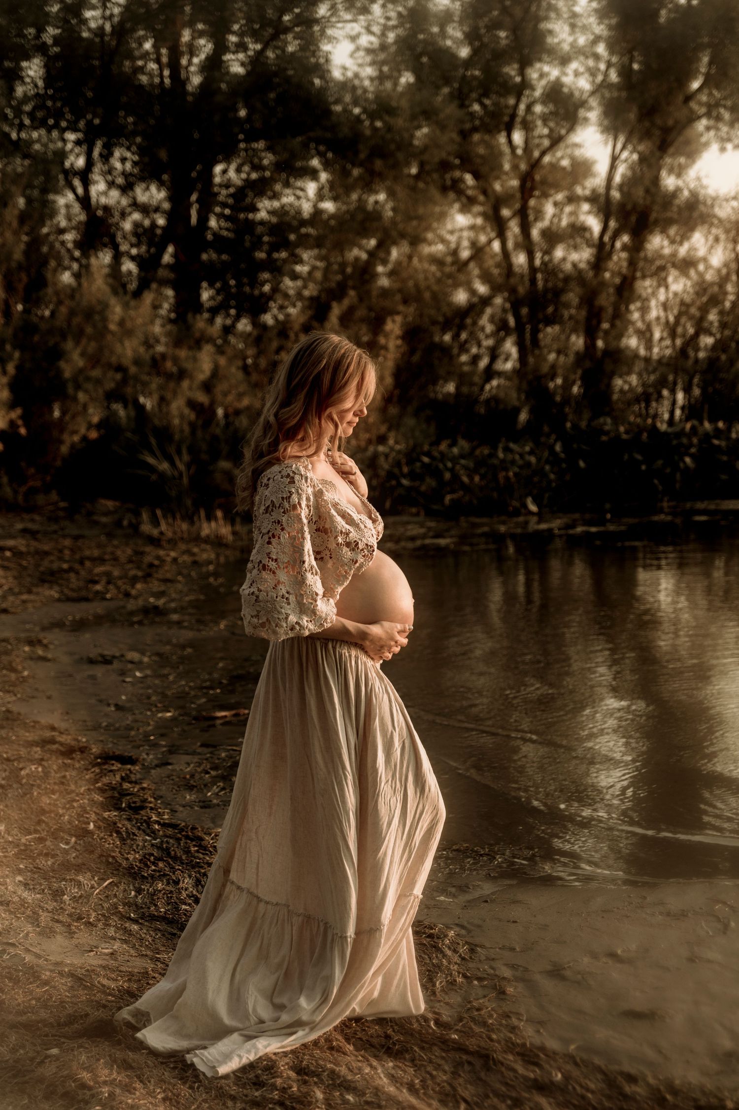 A pregnant woman in a flowing lace dress stands contemplatively by a peaceful lake at sunset.
