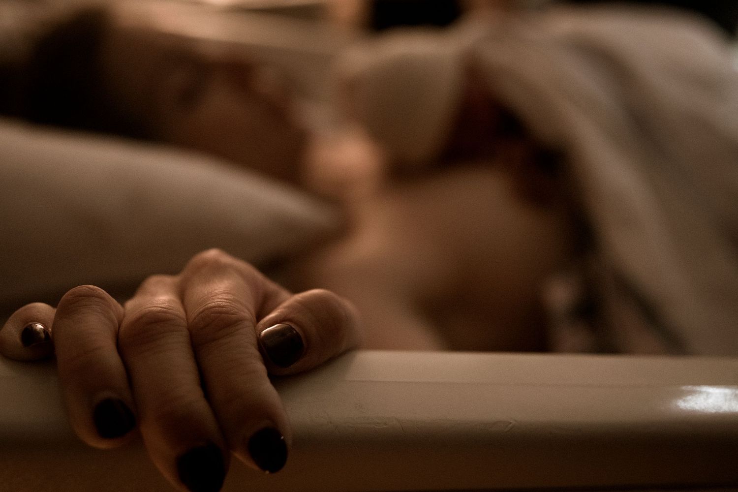 A hand with dark nail polish rests on white bedsheets in a dimly lit bedroom setting.