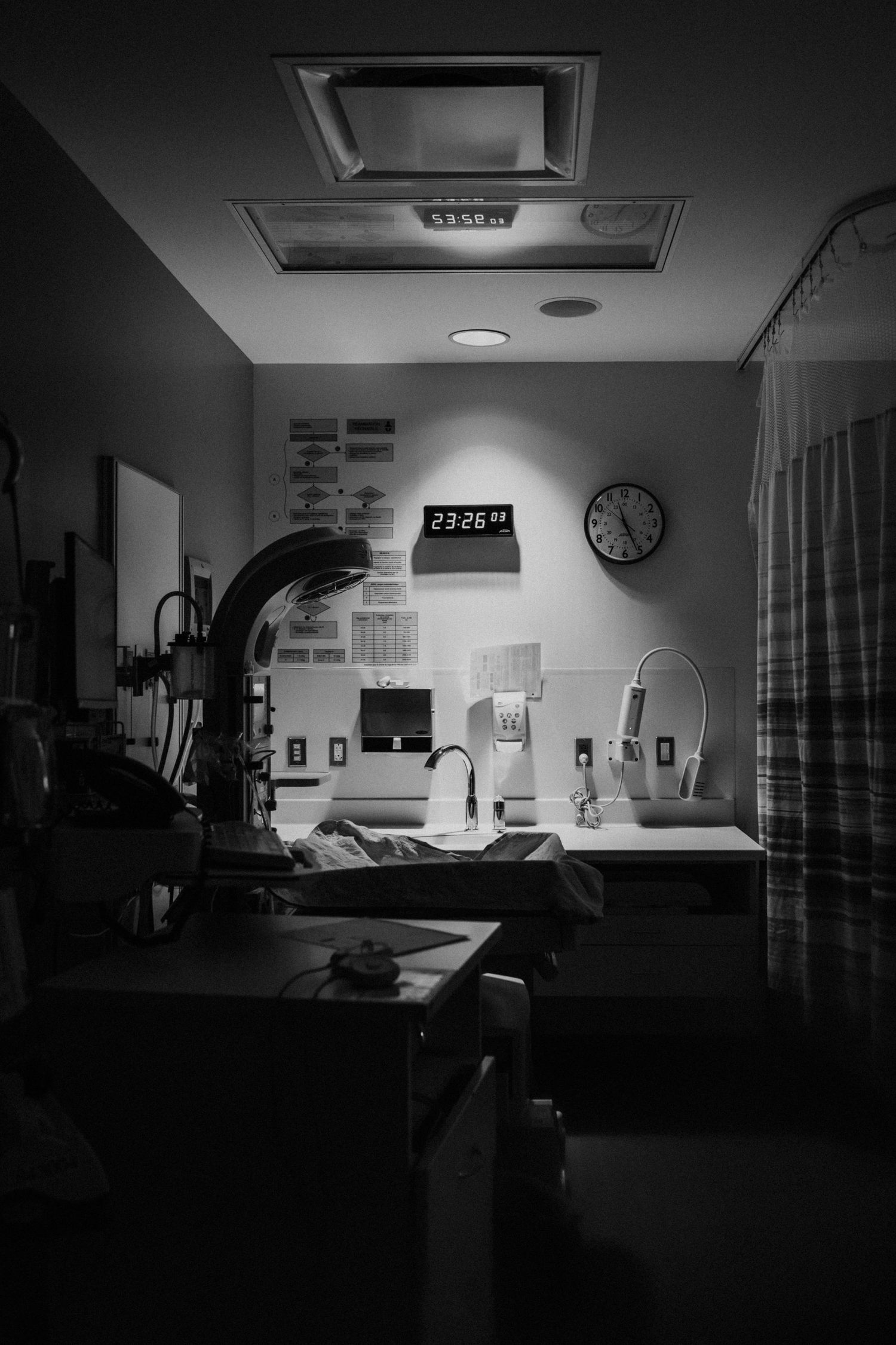 Dark black and white interior view of a dimly lit medical examination room with equipment and a wall clock.