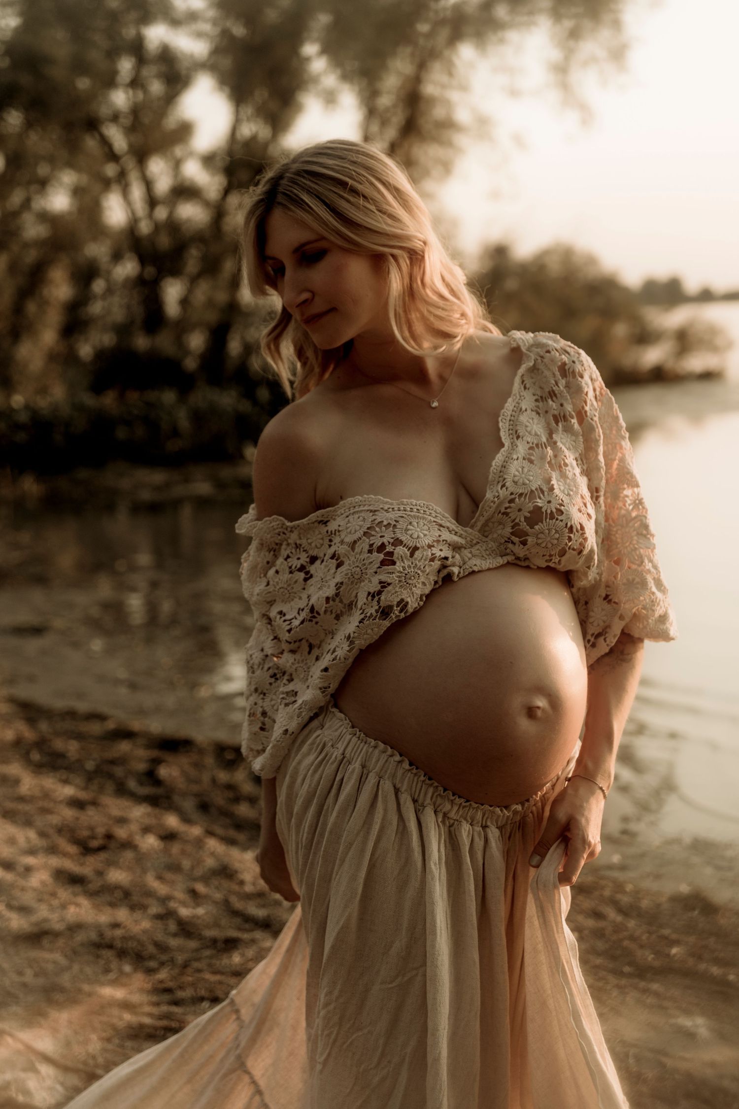 Artistic maternity photo of an expecting mother in a flowing lace dress standing by a lake at sunset.