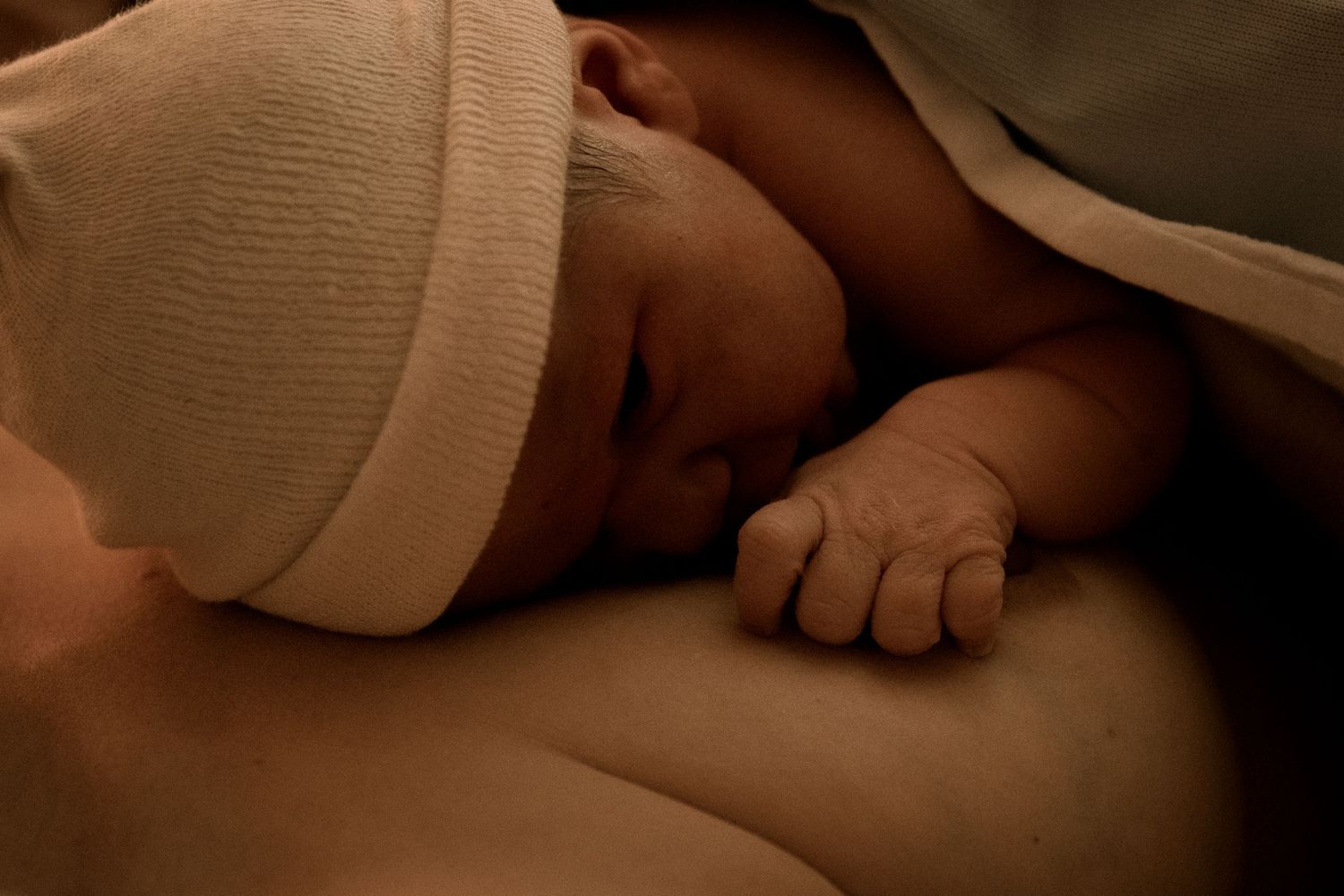 Newborn baby wearing white cap sleeping peacefully while skin-to-skin with parent in dim lighting.