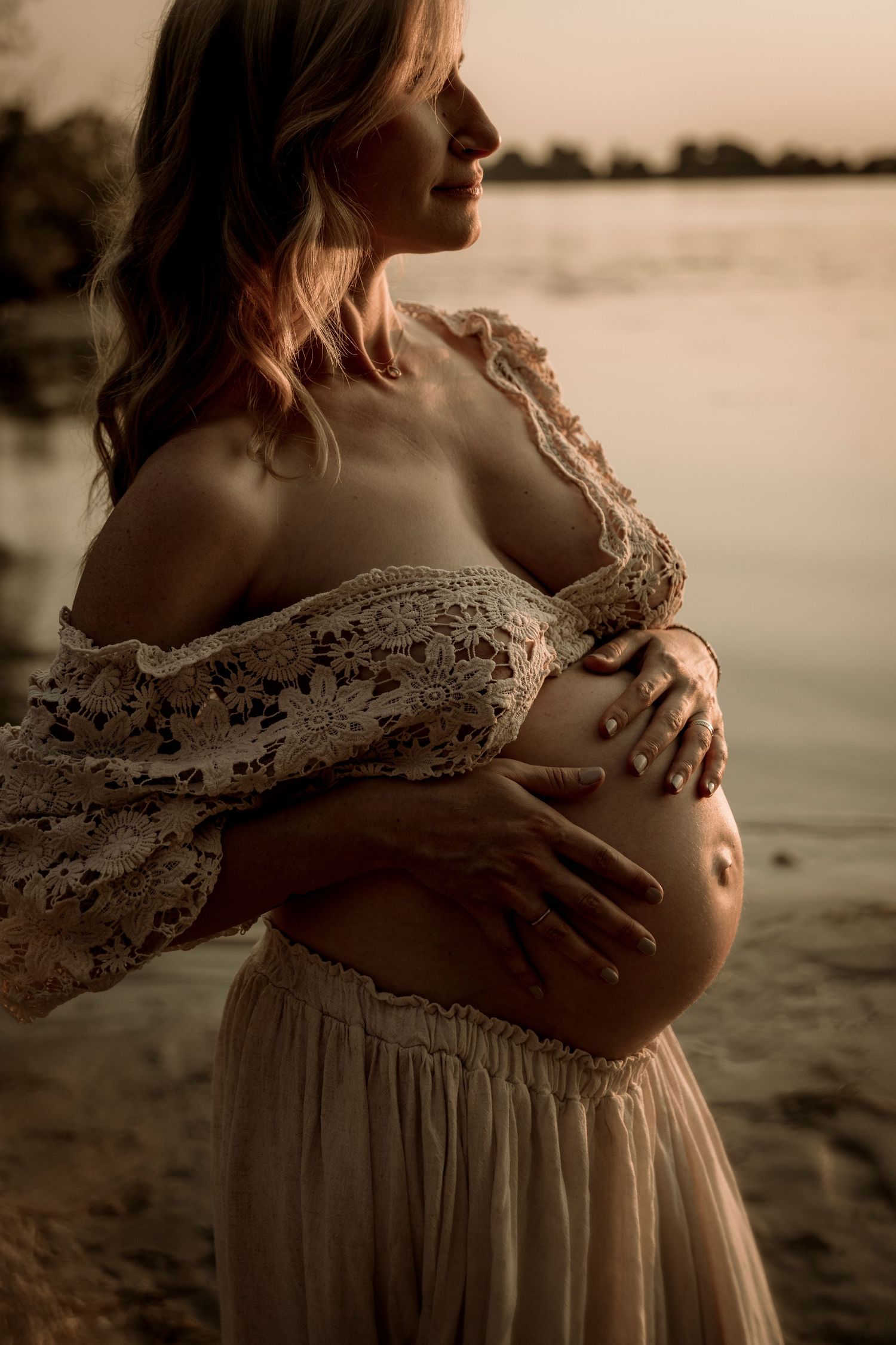 Pregnant woman in lace dress caresses her belly during sunset at a beach in warm sepia tones.