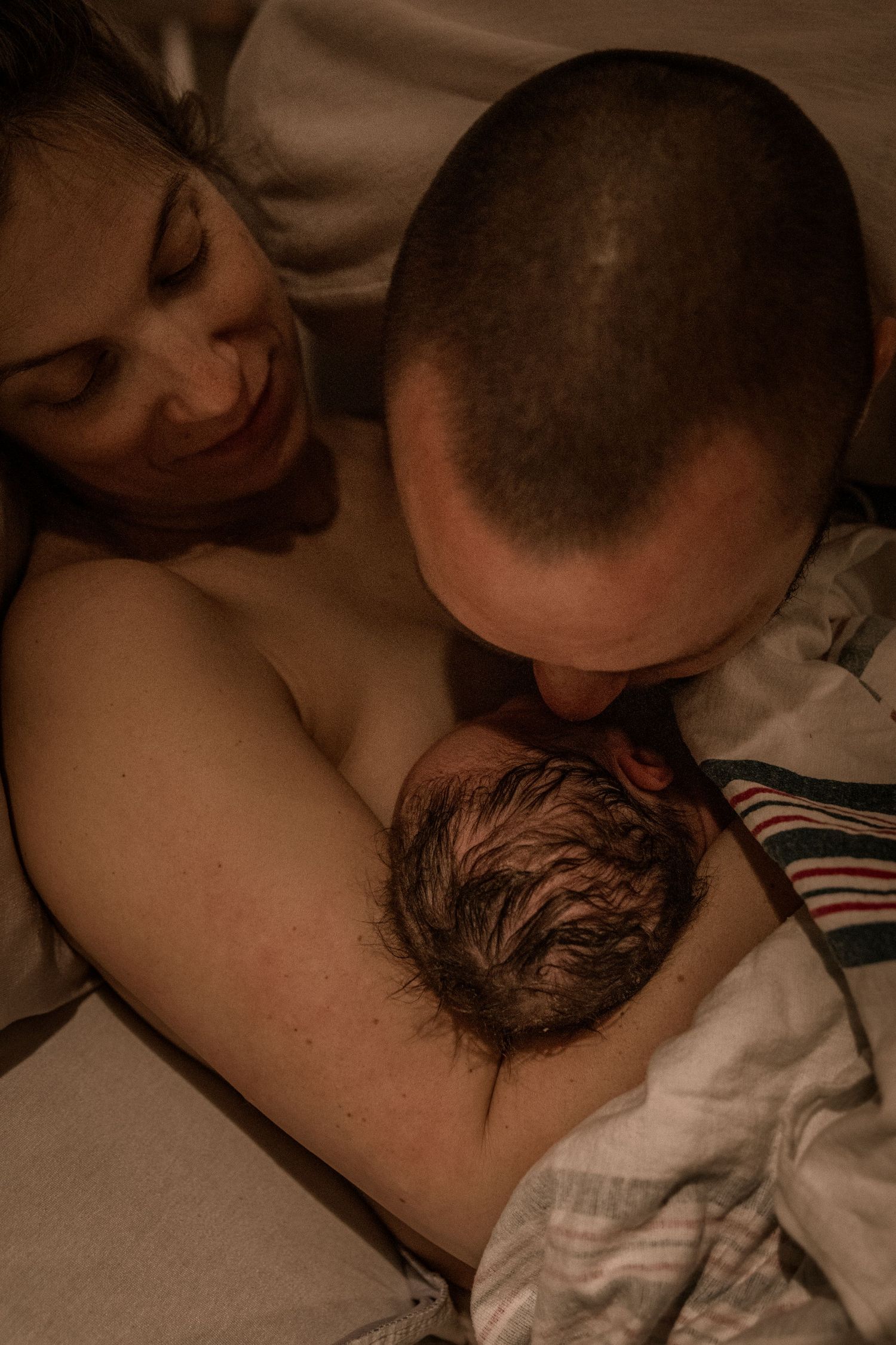 A tender moment as parents lovingly embrace their newborn baby in warm, sepia-toned lighting.