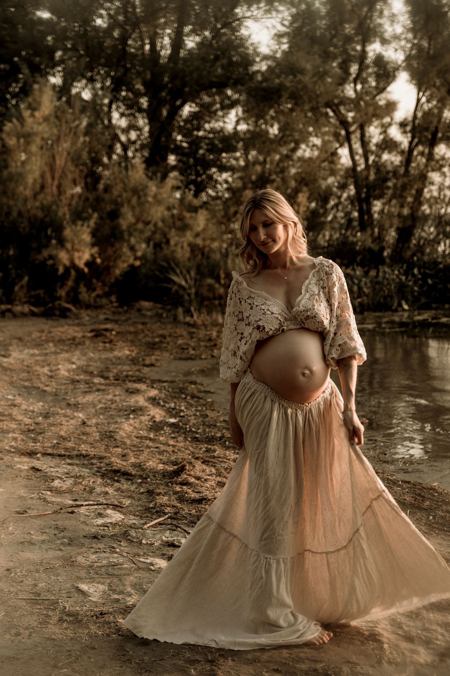 A pregnant woman in a flowing beige lace dress poses gracefully by a river at sunset in a dreamy outdoor setting.