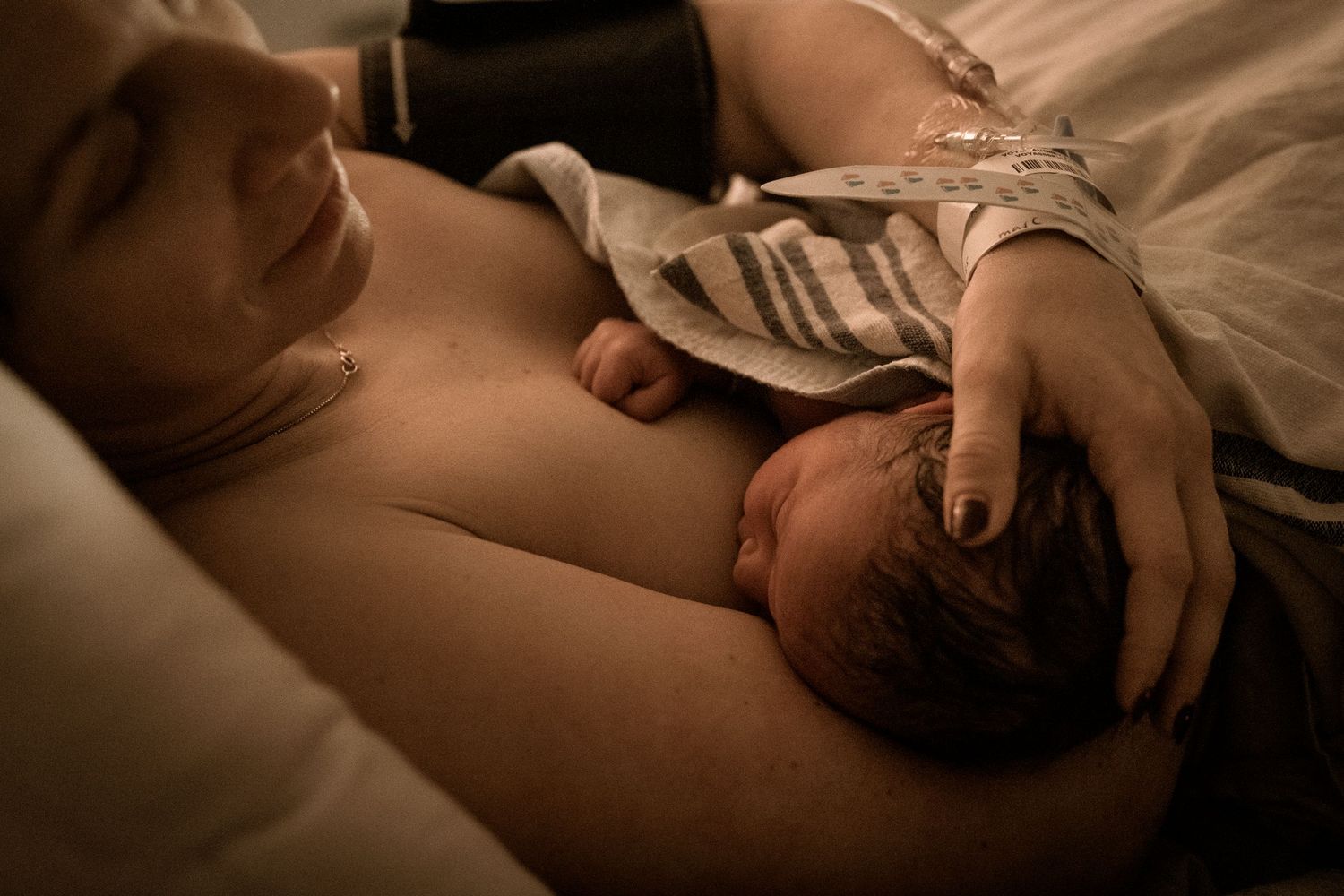 A newborn baby lies peacefully on a parent's bare chest in warm, intimate lighting during skin-to-skin bonding time.